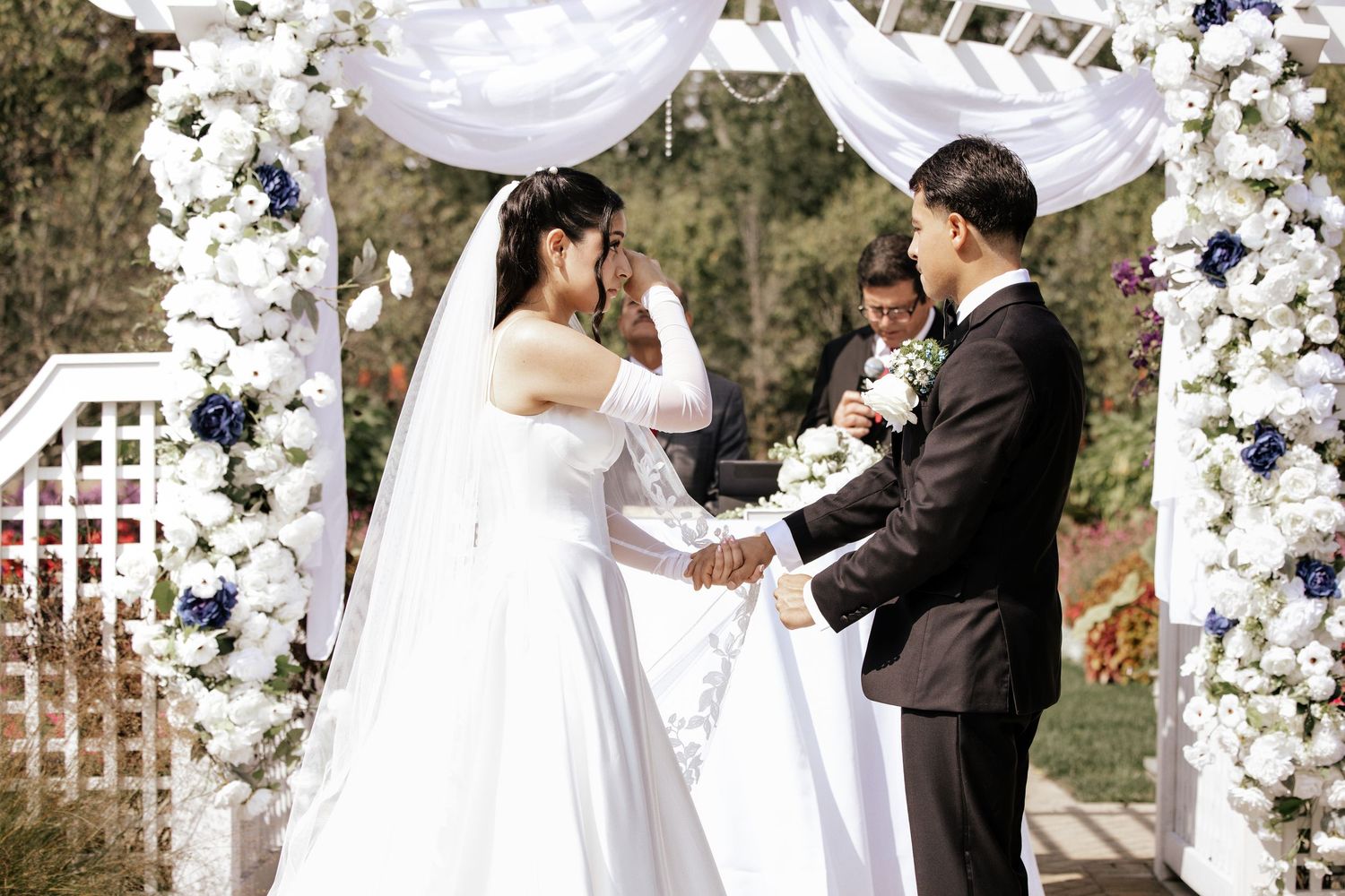 Bride and groom exchange vows under a white floral arch at an elegant outdoor wedding ceremony.
