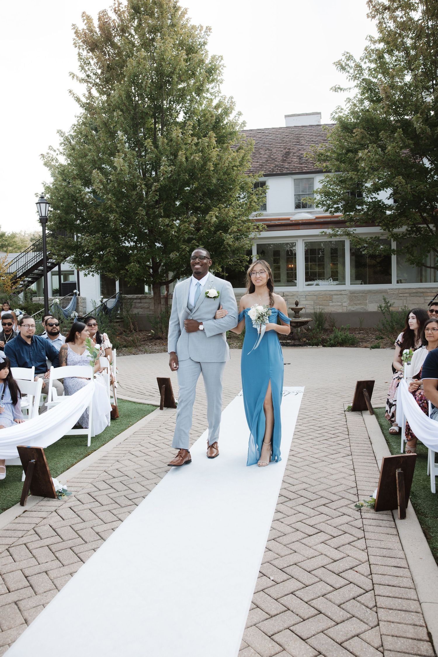 Groomsman in gray suit escorts bridesmaid in blue dress past seated wedding guests outdoors.