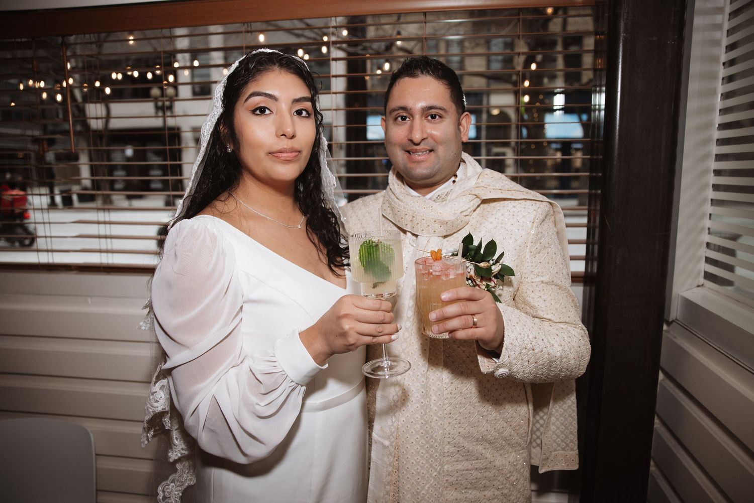 Bride and groom posing with cocktails at wedding reception, bride in white gown with veil, groom in traditional ivory sherwani.