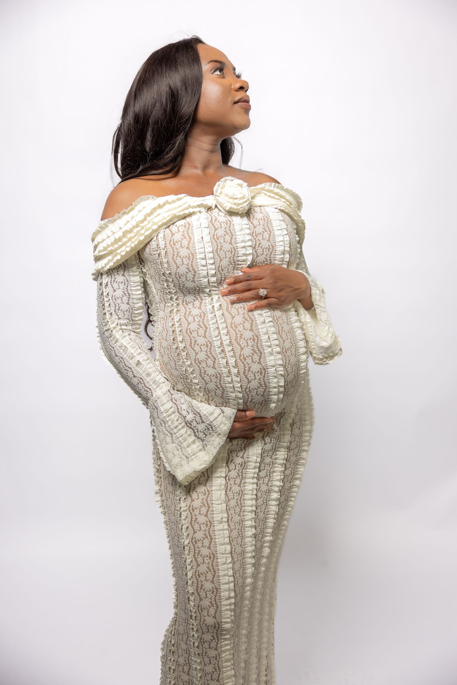 Pregnant Black woman in white lace off-shoulder gown gazing upward, holding baby bump.