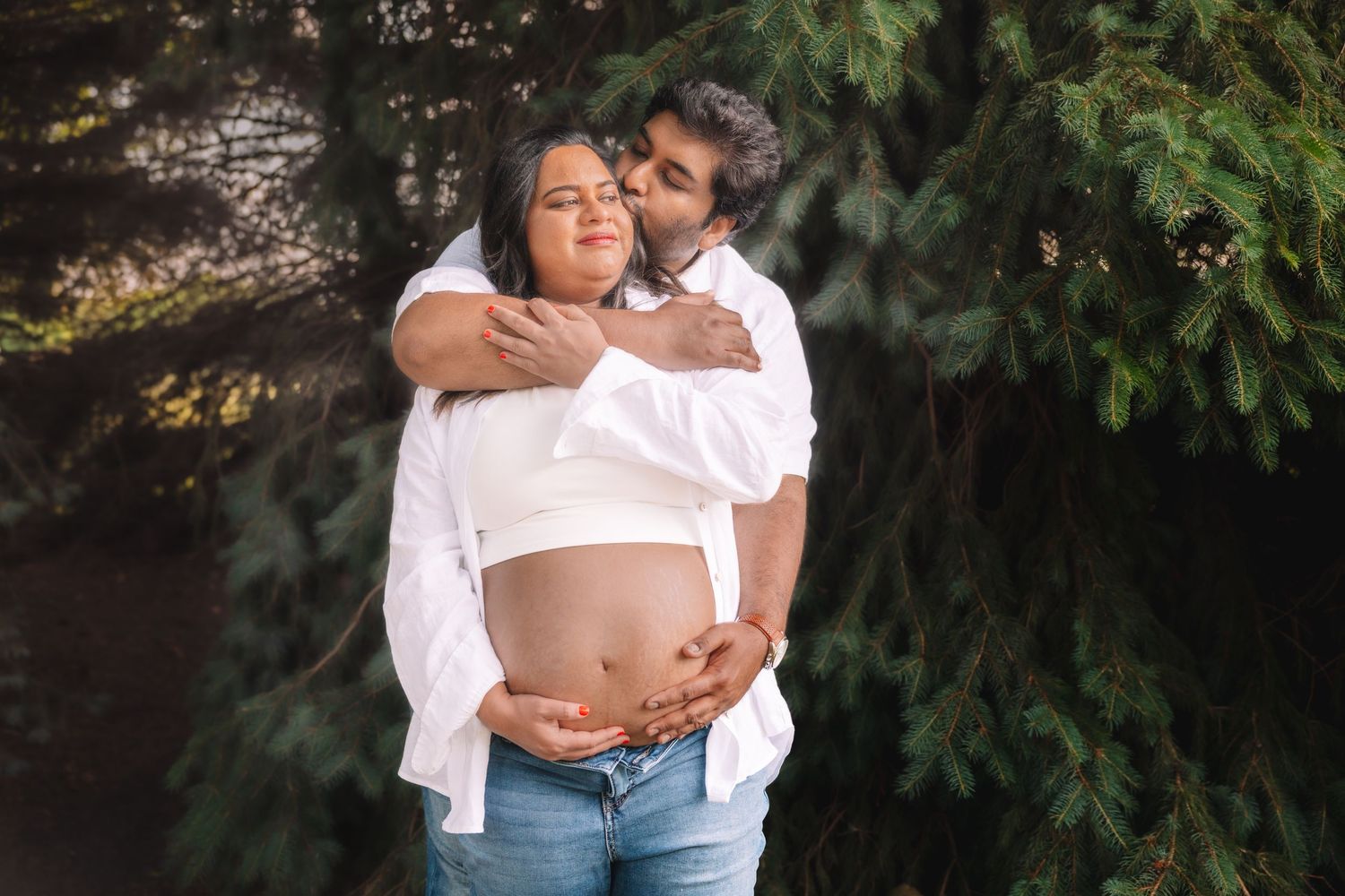 Couple embracing outdoors, man kissing pregnant woman's cheek while holding her baby bump near pine trees.