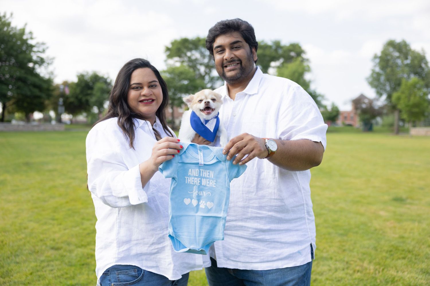 Couple in white holding a small dog wearing a blue baby onesie in a park.