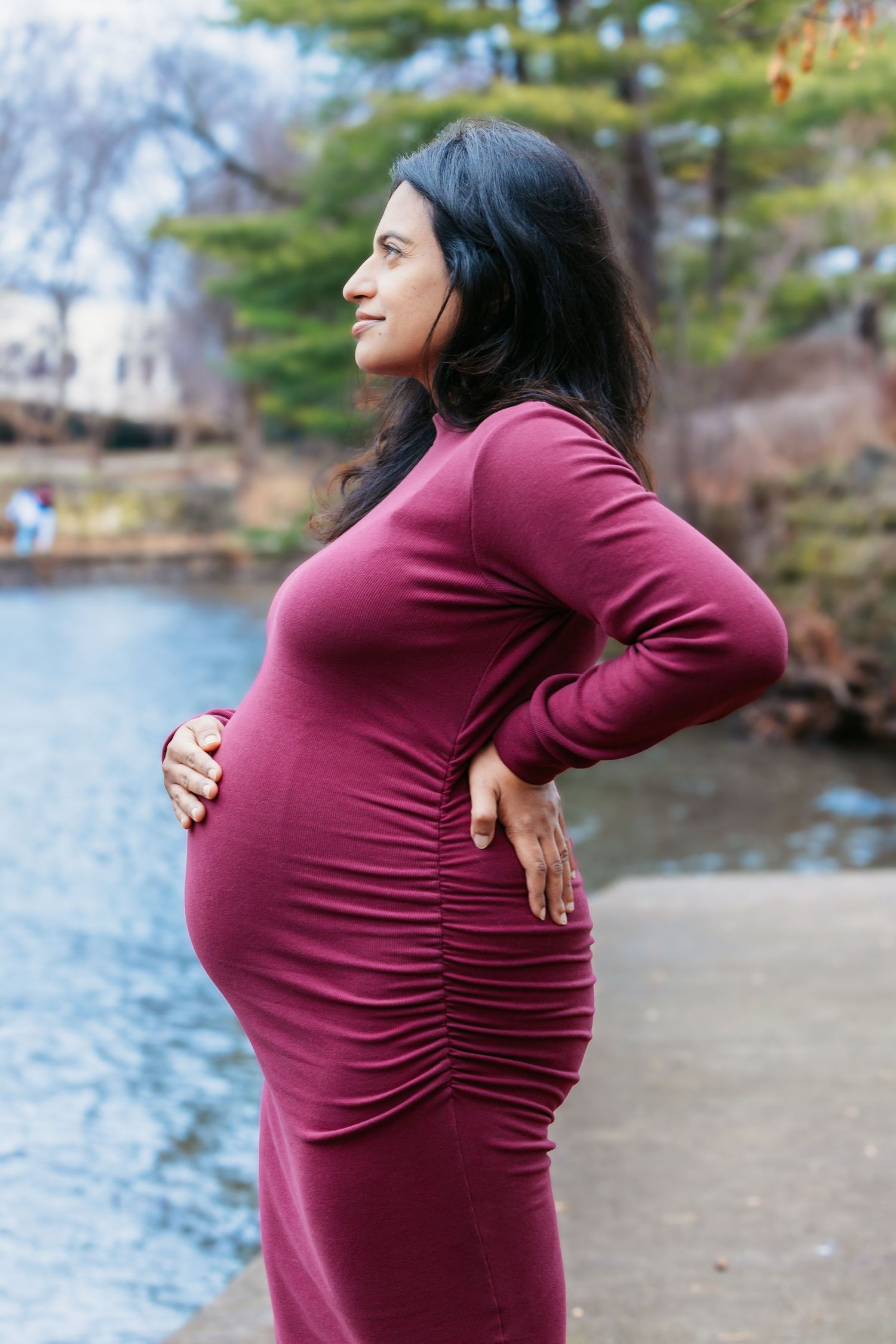 Pregnant woman in burgundy dress posing in profile, hand on hip near a peaceful park pond.