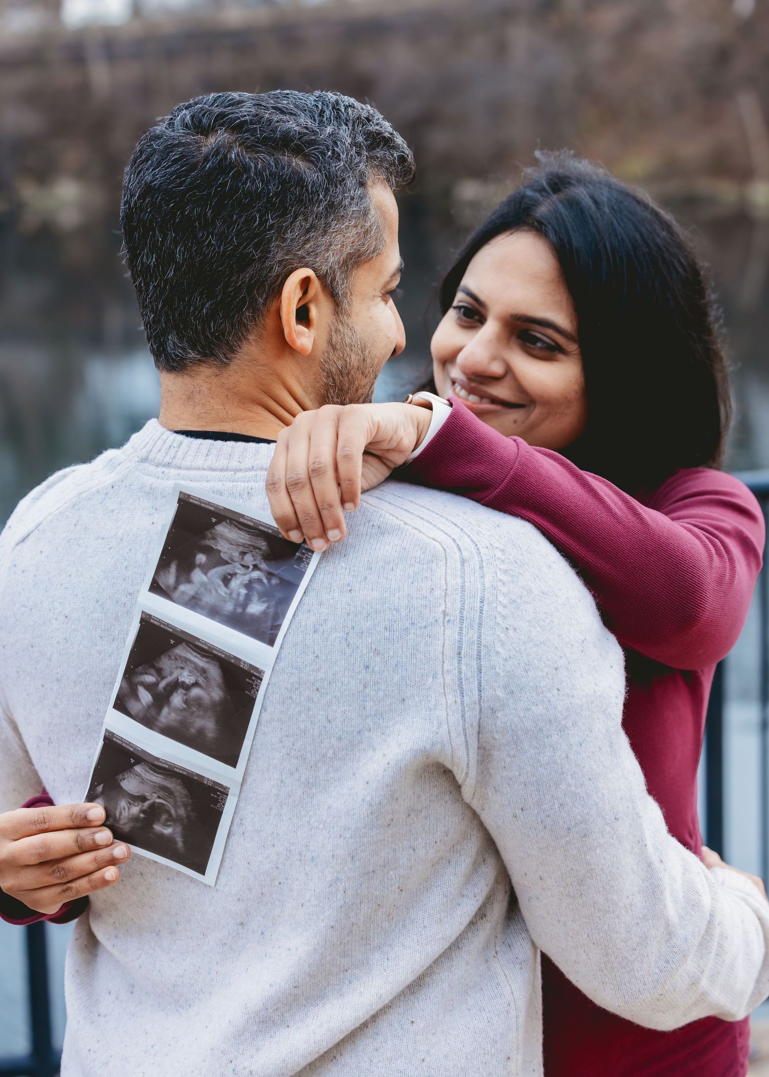 Happy couple hugging outside, man displaying ultrasound images while woman smiles warmly over his shoulder.