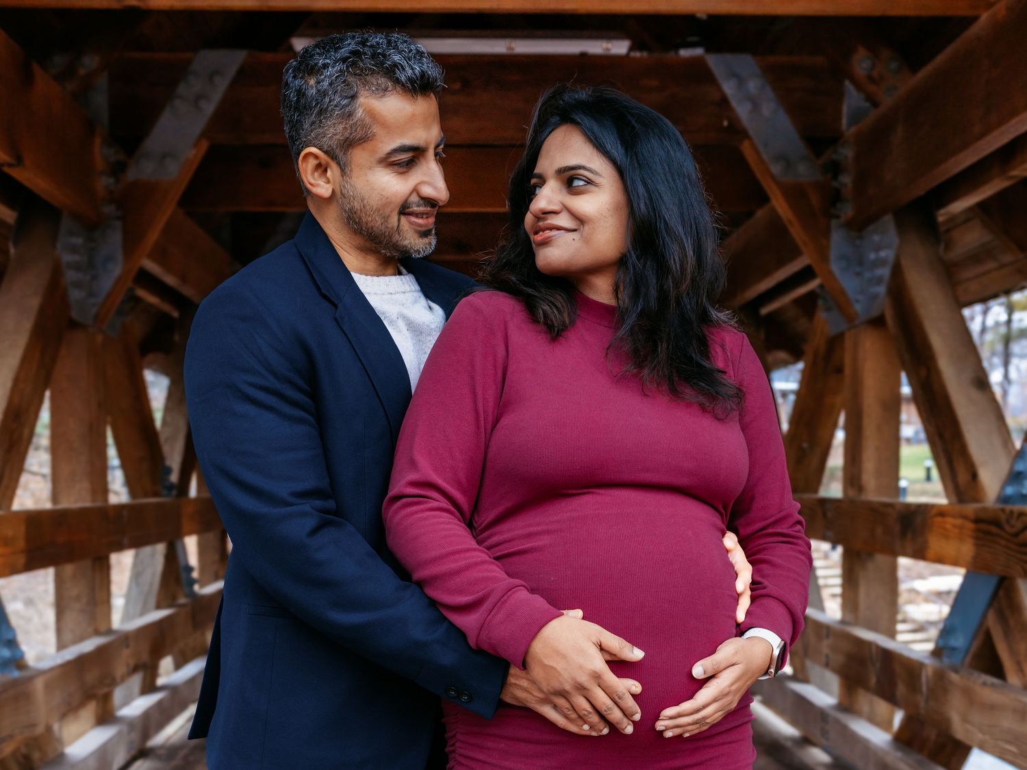 Expecting couple sharing a tender moment on a covered bridge, man embracing pregnant woman in burgundy dress.