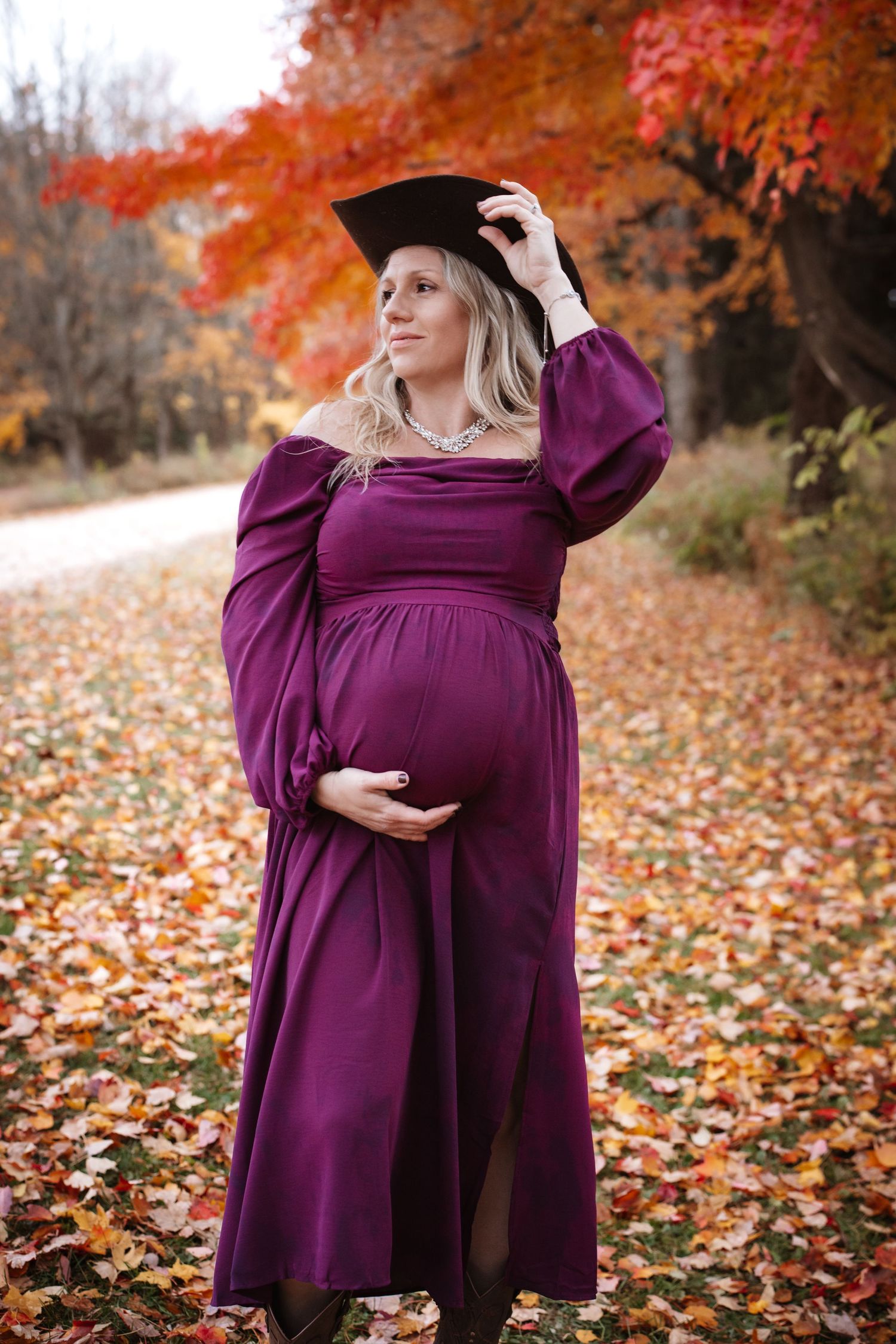 Elegant pregnant woman in purple gown wearing graduation cap standing among colorful fall leaves outdoors.