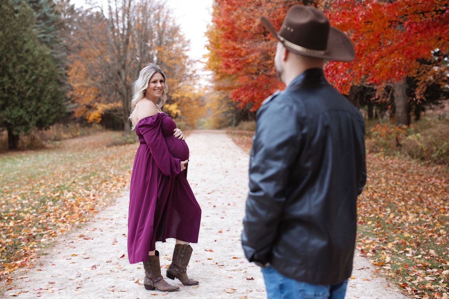 Expecting woman in burgundy gown smiles at her partner on a colorful fall foliage path.