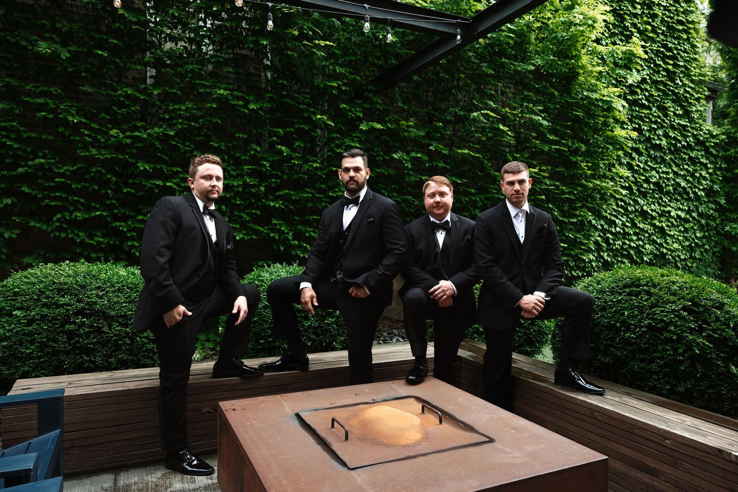 Four groomsmen in black tuxedos leaning near an outdoor fire pit with ivy wall behind.