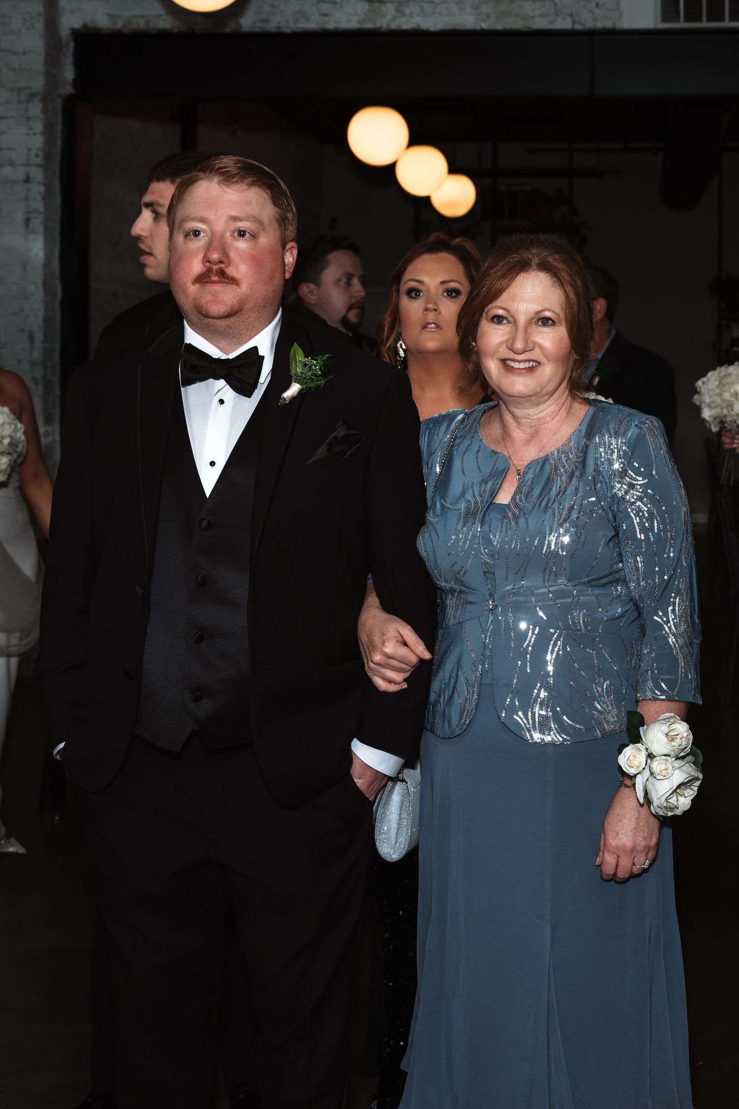 Groom in tuxedo escorting mother in blue gown during wedding processional ceremony.