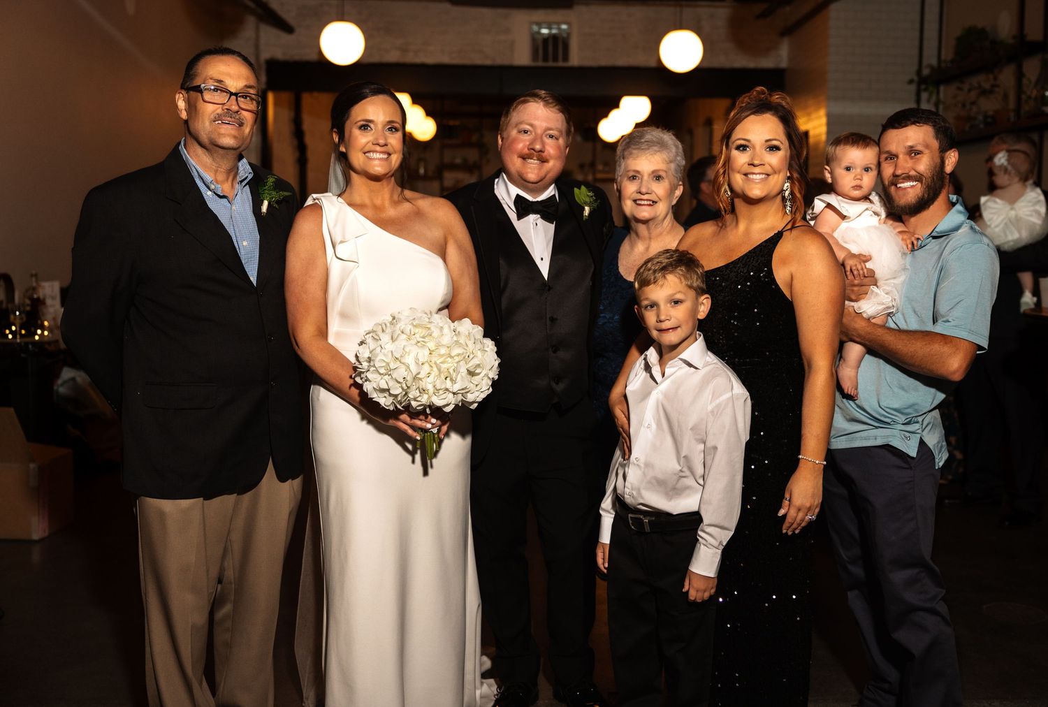 Wedding party group photo with bride, groom, and six family members including children at indoor reception venue.
