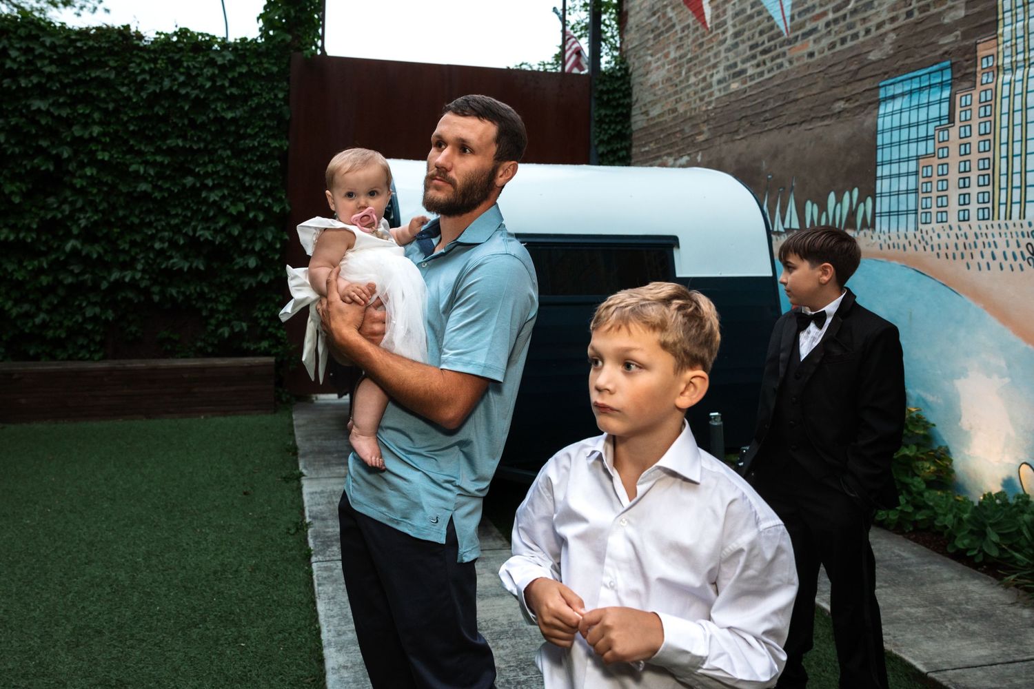 A man holds a baby in white while a young boy stands nearby at an outdoor wedding venue.
