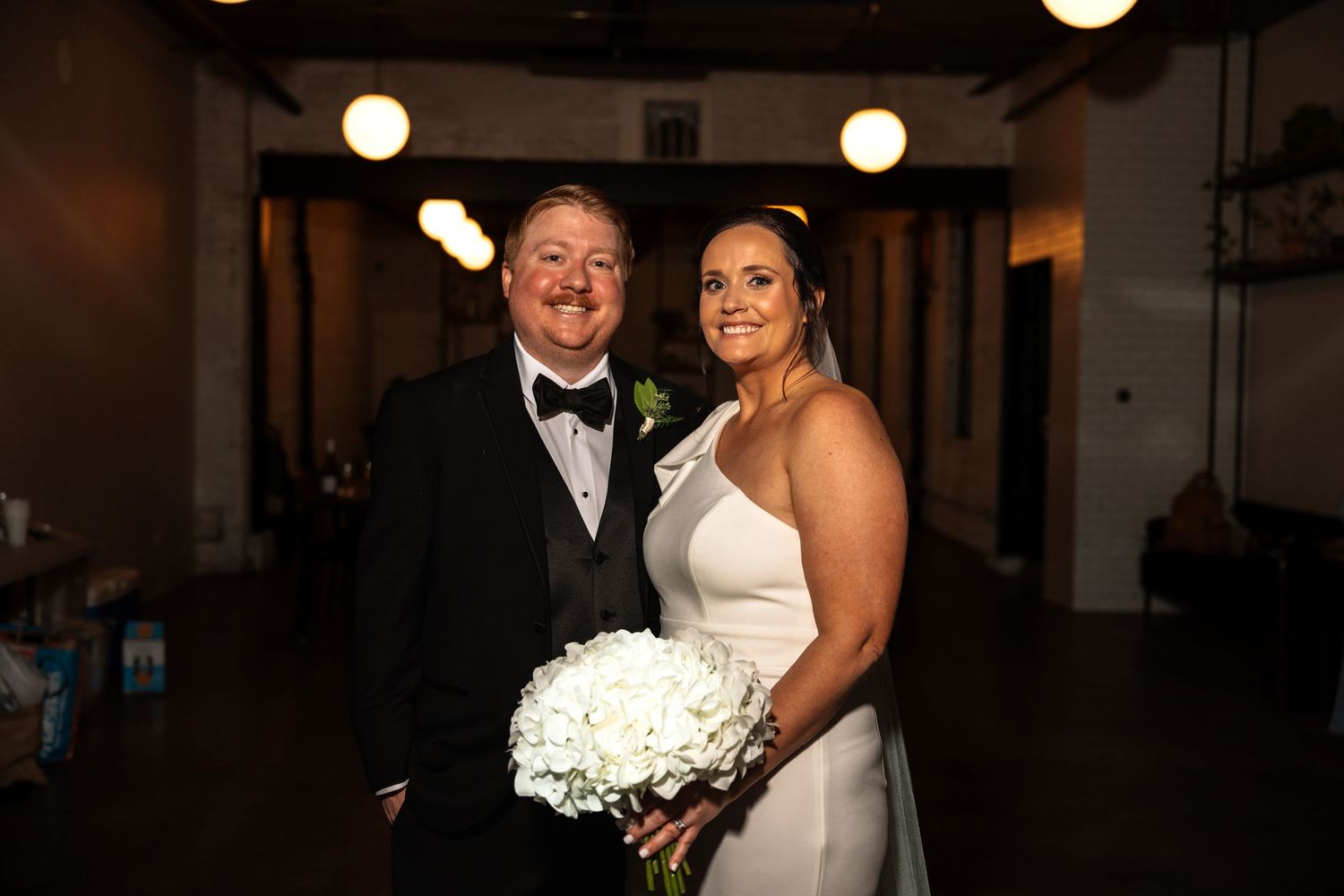 Smiling bride and groom pose together, bride holding white hydrangea bouquet at wedding reception.