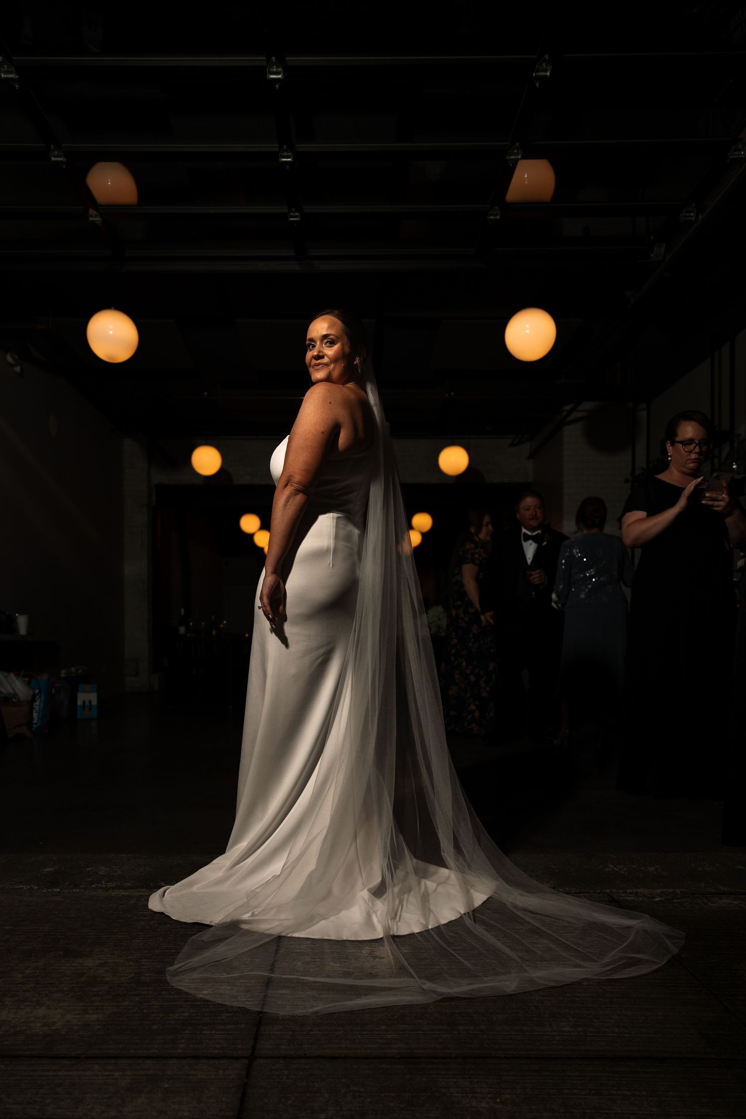 Stunning bride in white fitted gown with long sheer veil poses in dramatic lighting in industrial event space.
