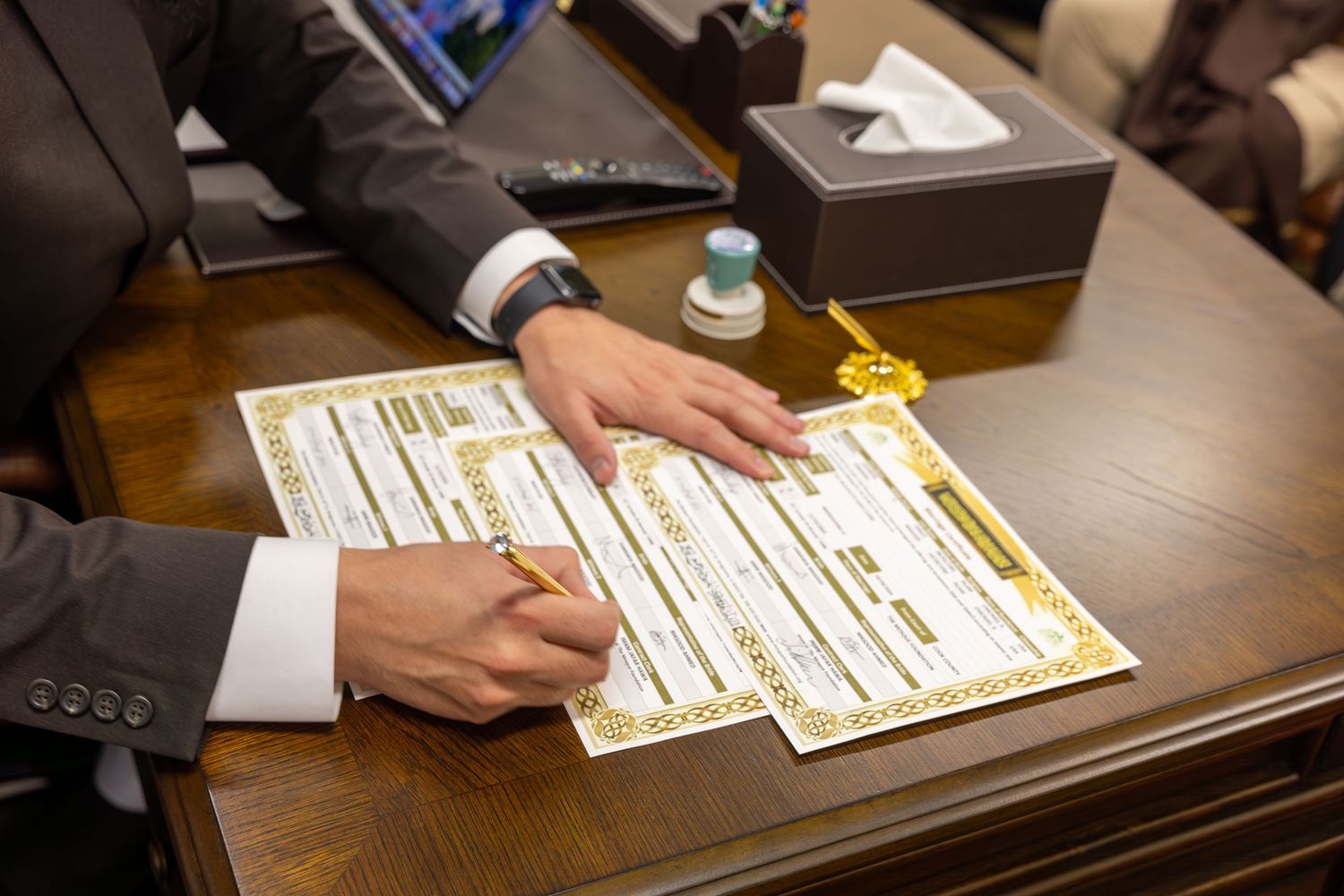 Person in gray suit signing official Islamic marriage certificate documents at a wooden desk.