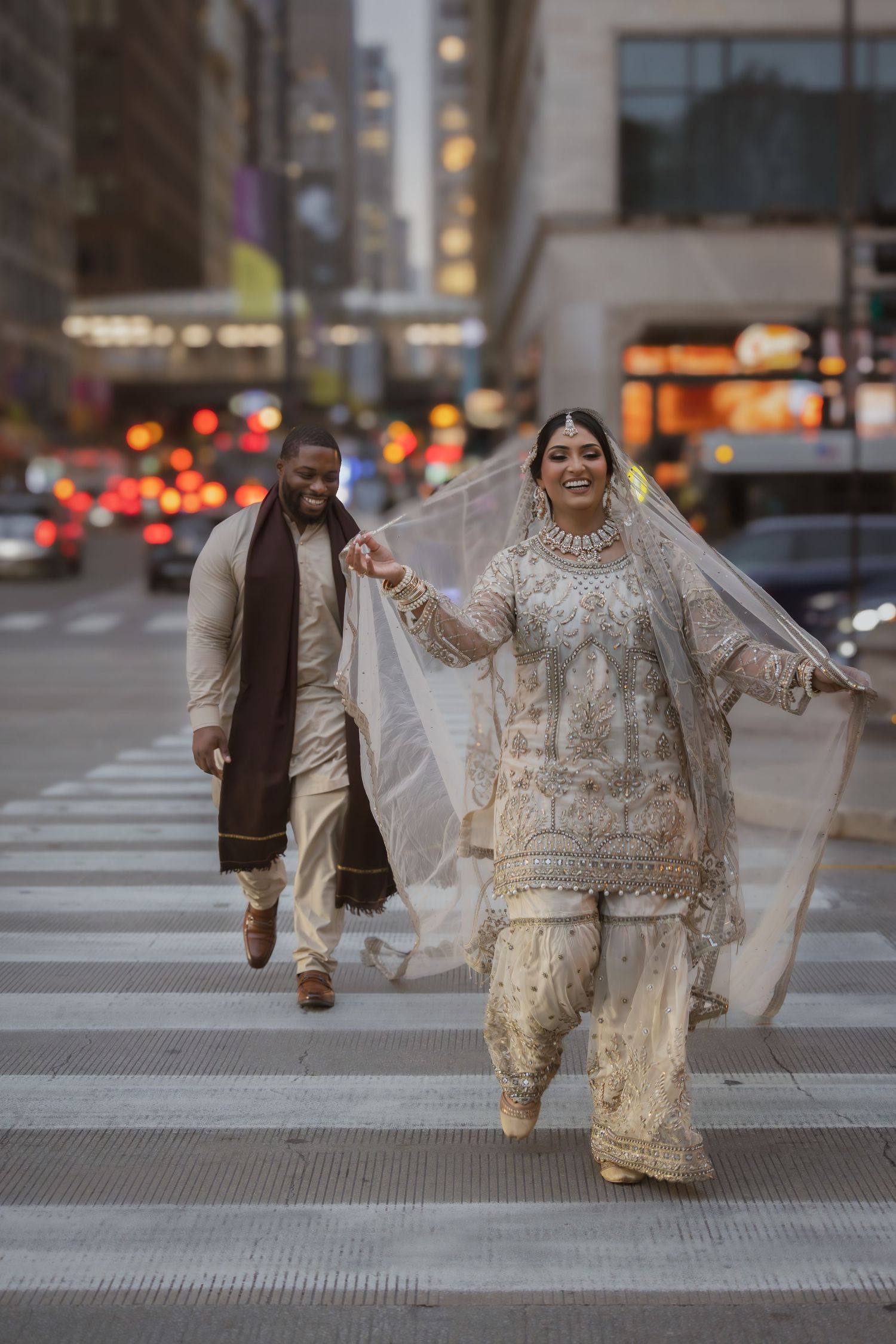 Joyful South Asian bride in white embroidered bridal outfit leads groom across a city crosswalk at dusk.