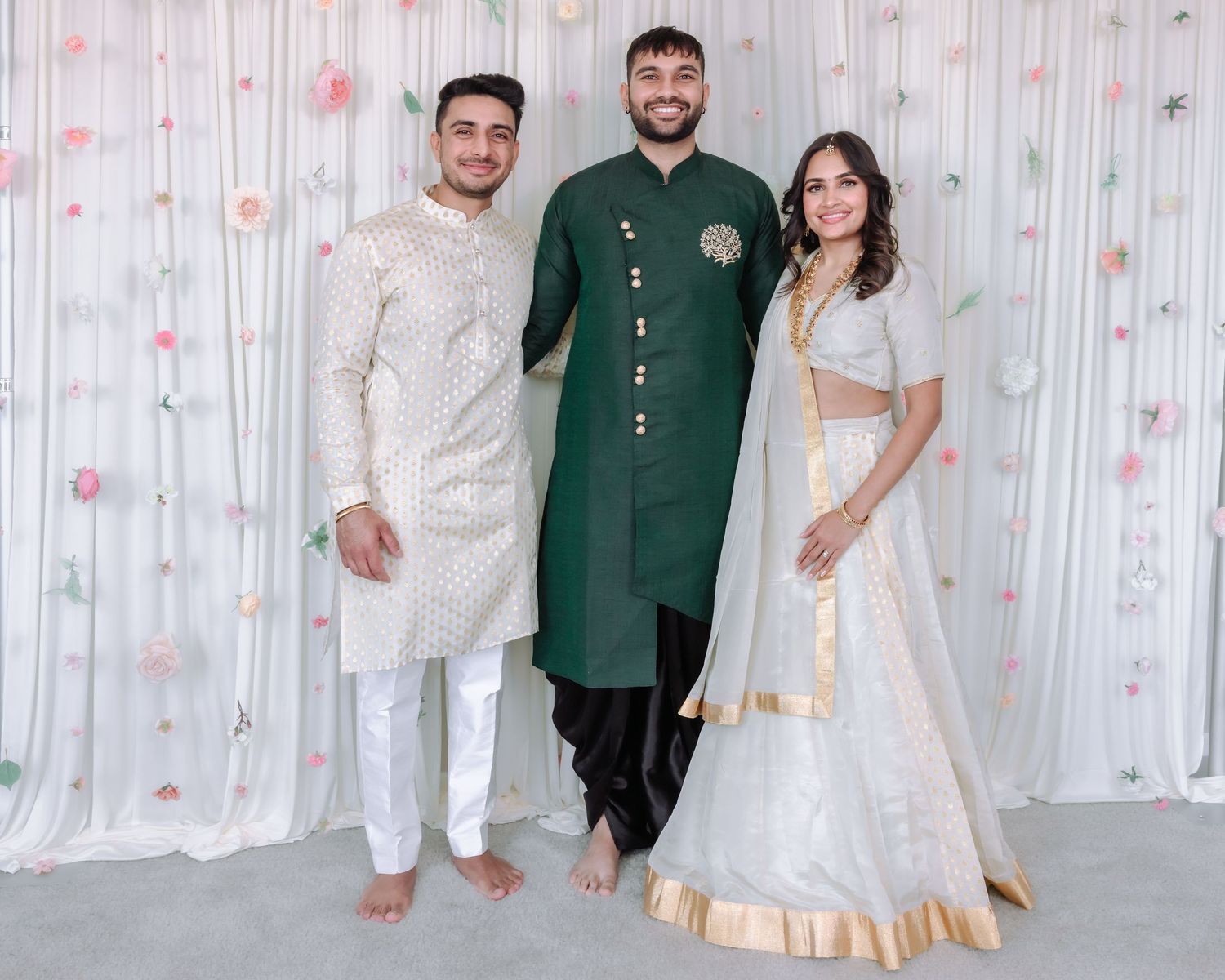 Three friends posing at Indian engagement, man in green sherwani flanked by couple in white.