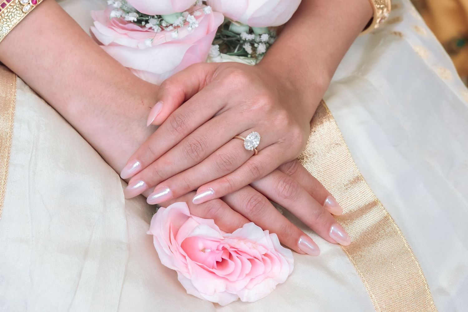 Close-up of bride's hands with diamond engagement ring and pink rose on white saree.