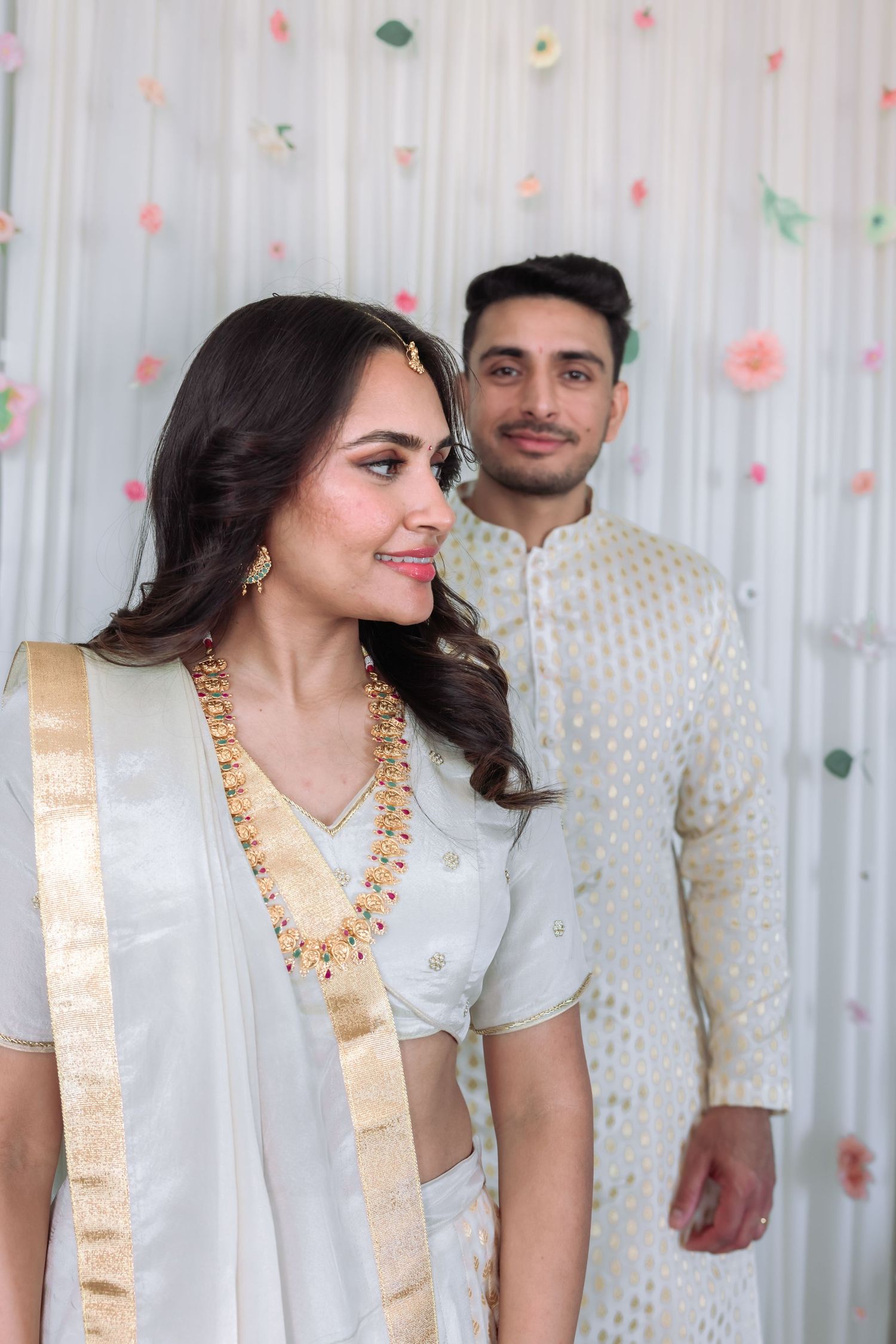 Indian couple in white and gold traditional attire posing elegantly before a floral backdrop.