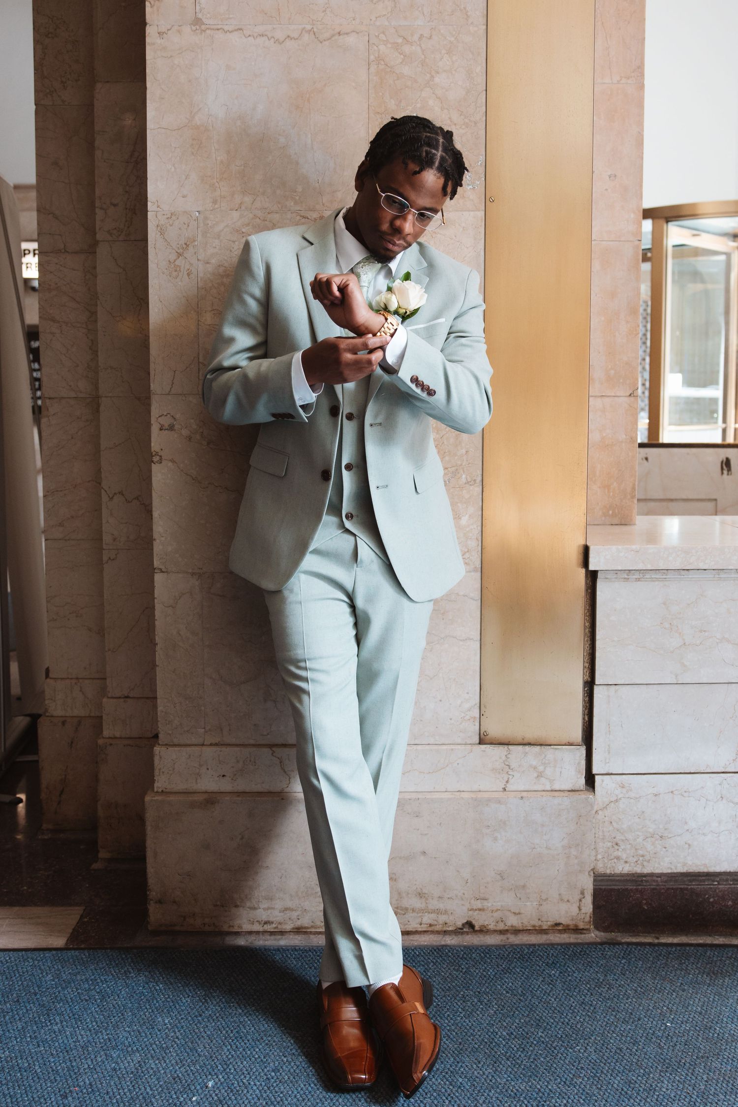 Groom in light gray three-piece suit adjusting boutonniere while leaning against marble pillar.