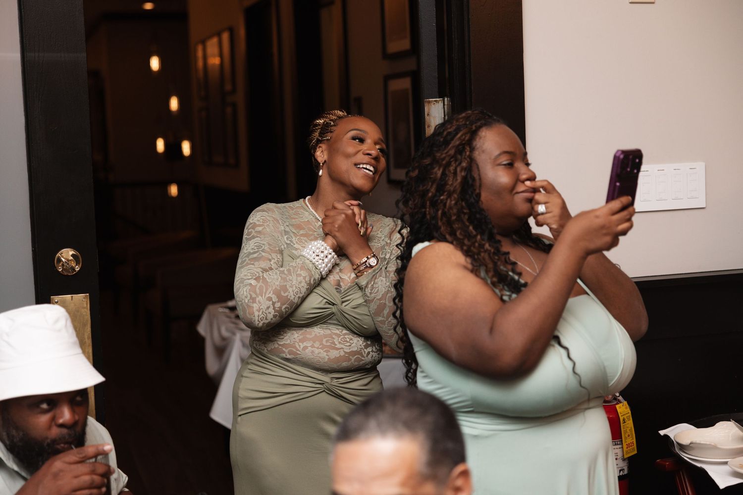 Two women in sage green dresses laugh and record video on smartphone during wedding reception celebration.