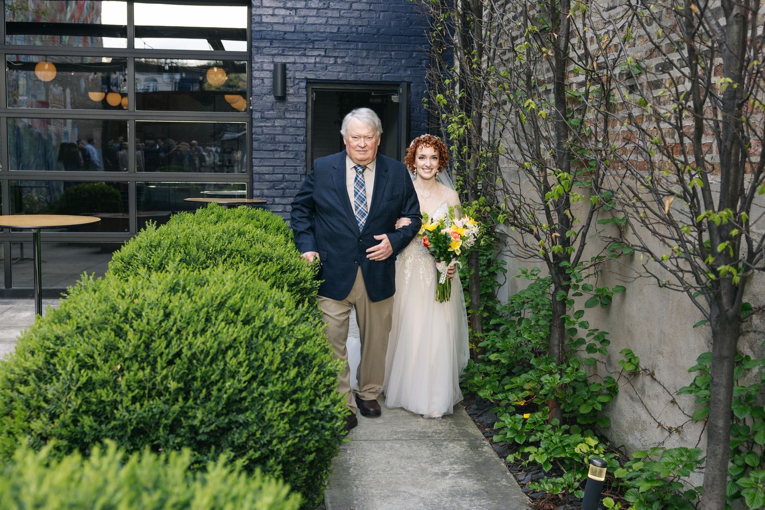 Bride in white gown holding colorful bouquet walks with older man in navy suit along garden path at outdoor wedding venue.