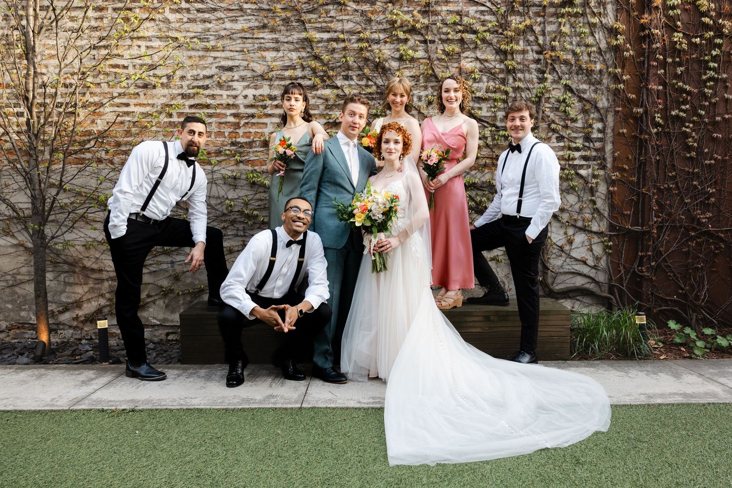 Wedding party posing outdoors against ivy wall, bride in white gown with bouquet, groom in teal suit, bridesmaids and groomsmen.