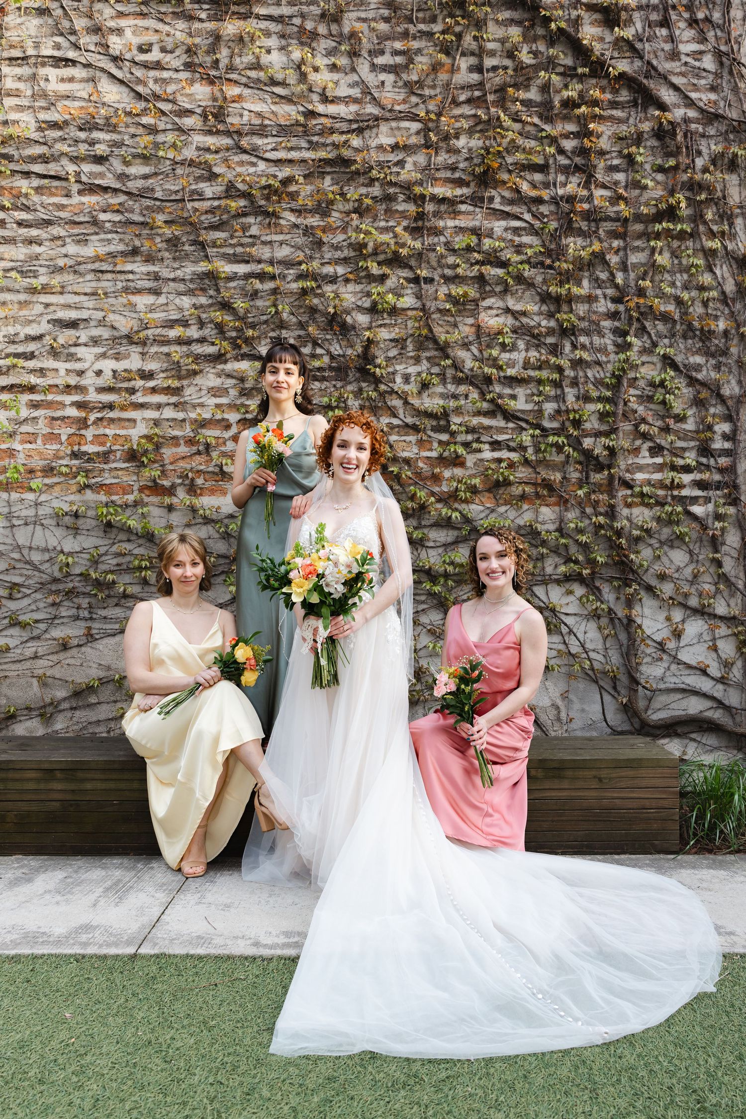 Bride in white gown poses with three bridesmaids in colorful dresses holding bouquets against ivy-covered brick wall.