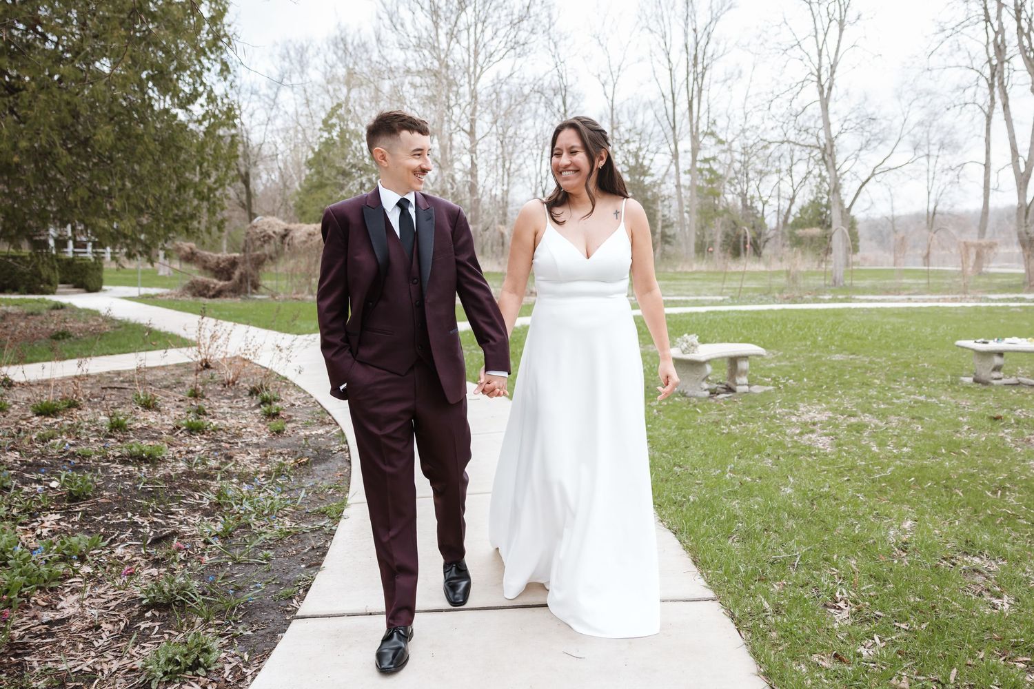 A smiling bride and groom walk hand in hand on a garden path, she in a white gown, he in a burgundy suit.