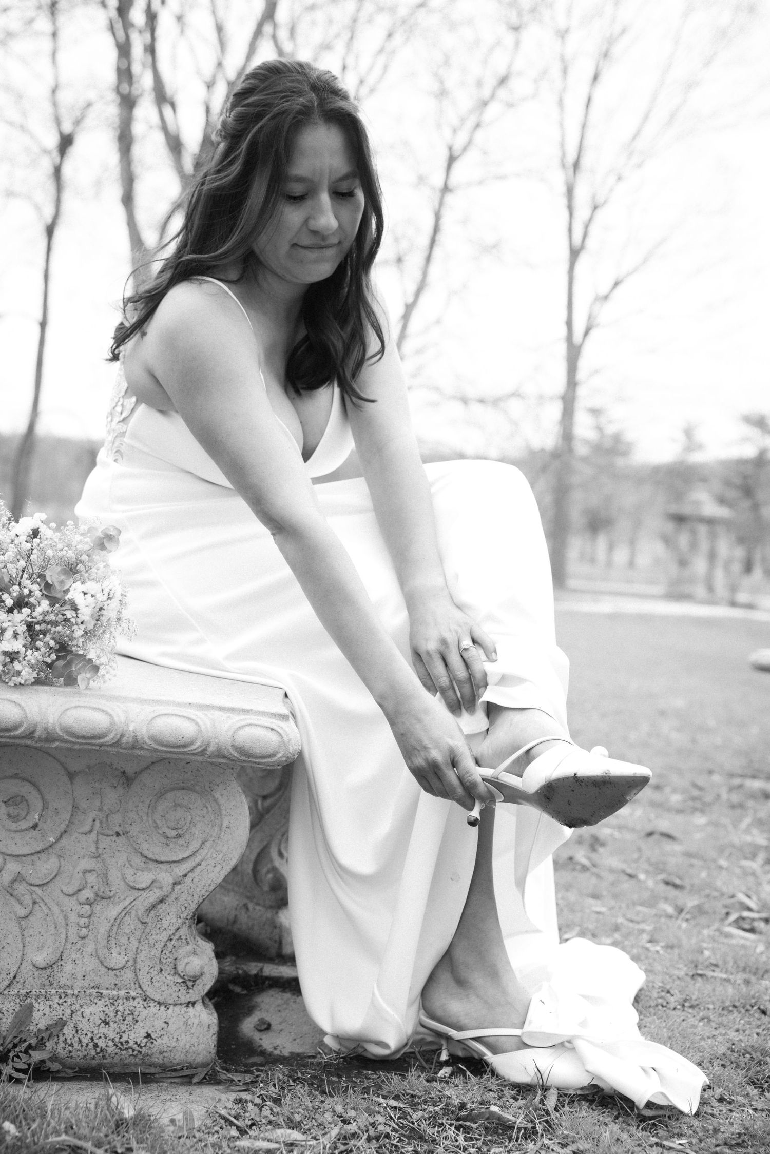 Black and white photo of a bride in white gown adjusting her shoe while seated on an ornate stone bench outdoors.
