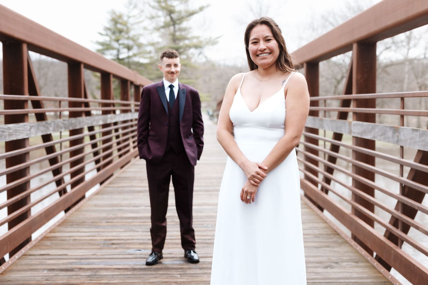 Bride in white dress and groom in burgundy suit posing on a wooden bridge outdoors.