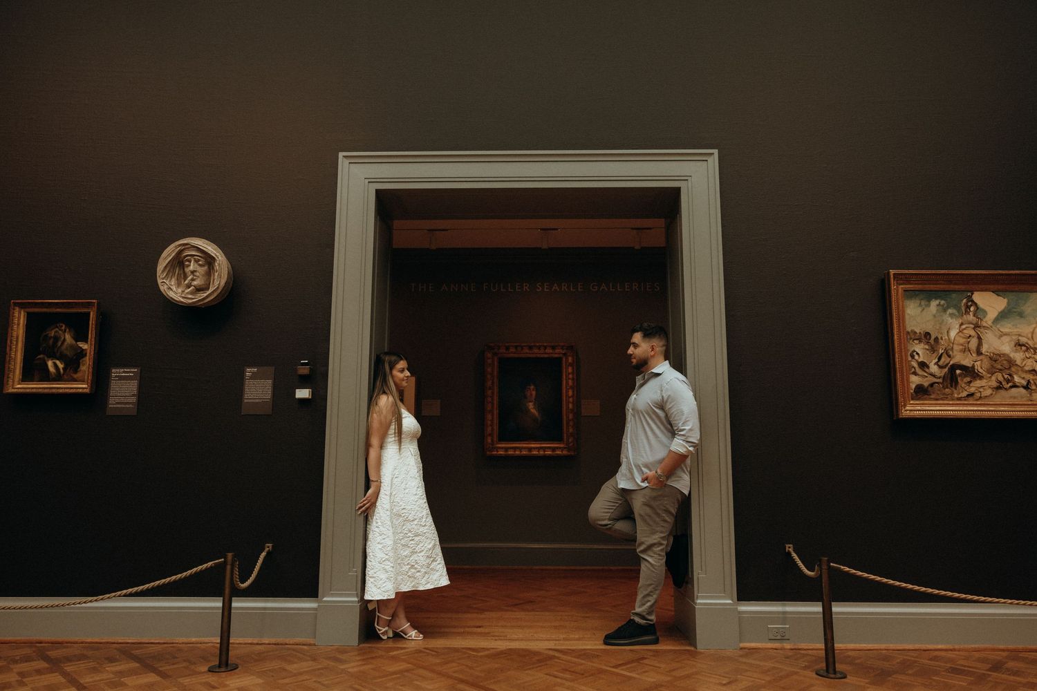 Couple standing apart in elegant art museum gallery doorway, surrounded by framed paintings on dark walls.