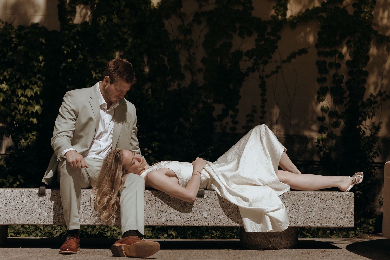 Bride reclines on outdoor bench while groom gazes at her lovingly in sunny garden setting.