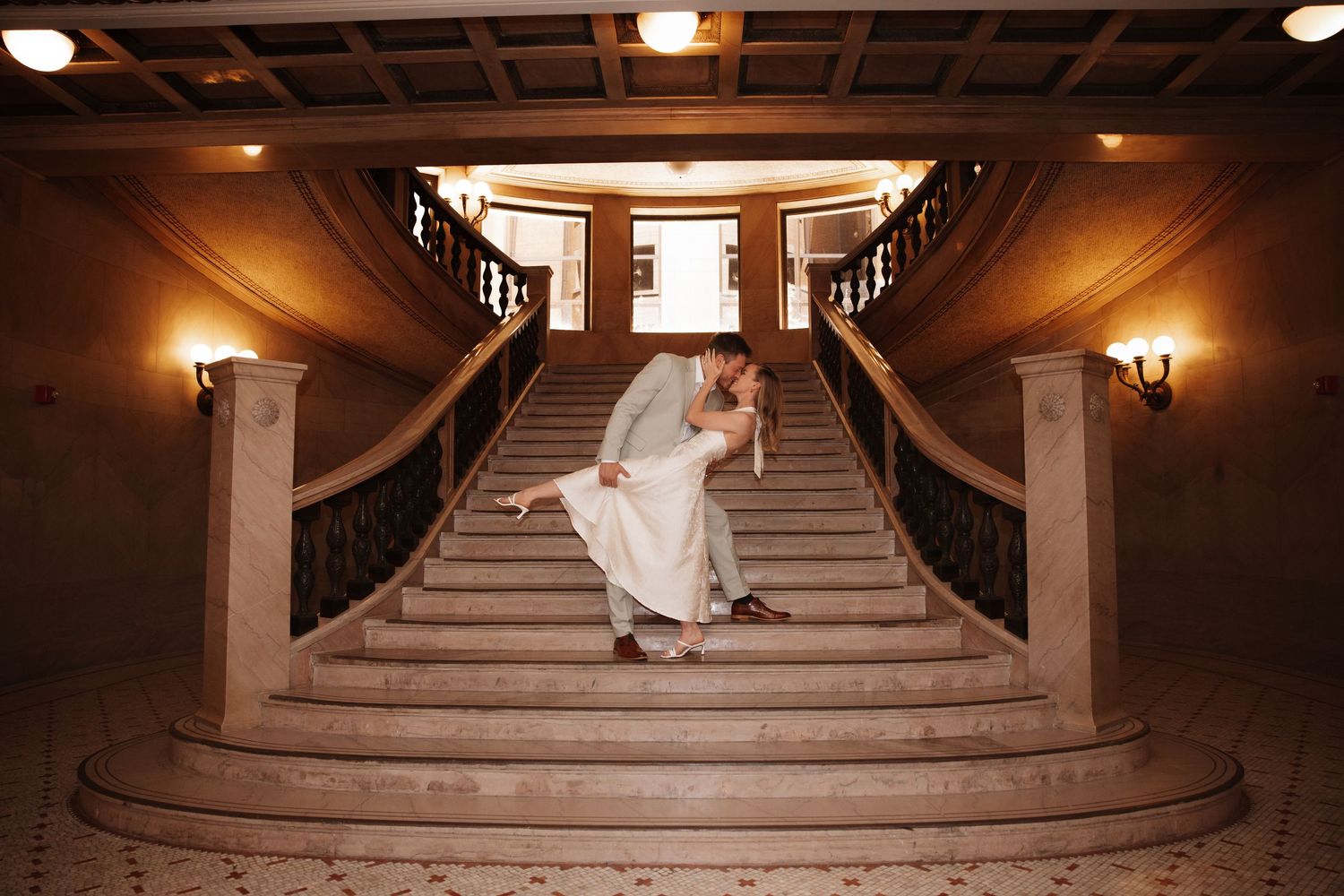 Bride and groom share a romantic dip kiss on grand marble staircase in elegant wedding venue.