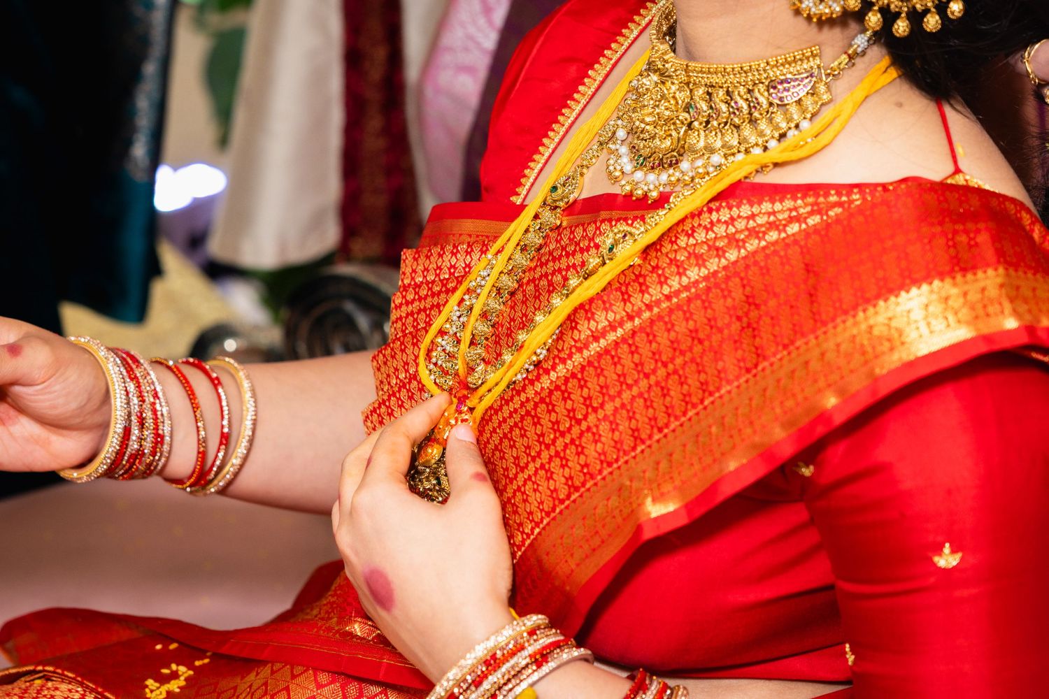 Close-up of Indian bride in red silk saree with gold bridal jewelry being adorned during wedding ceremony.