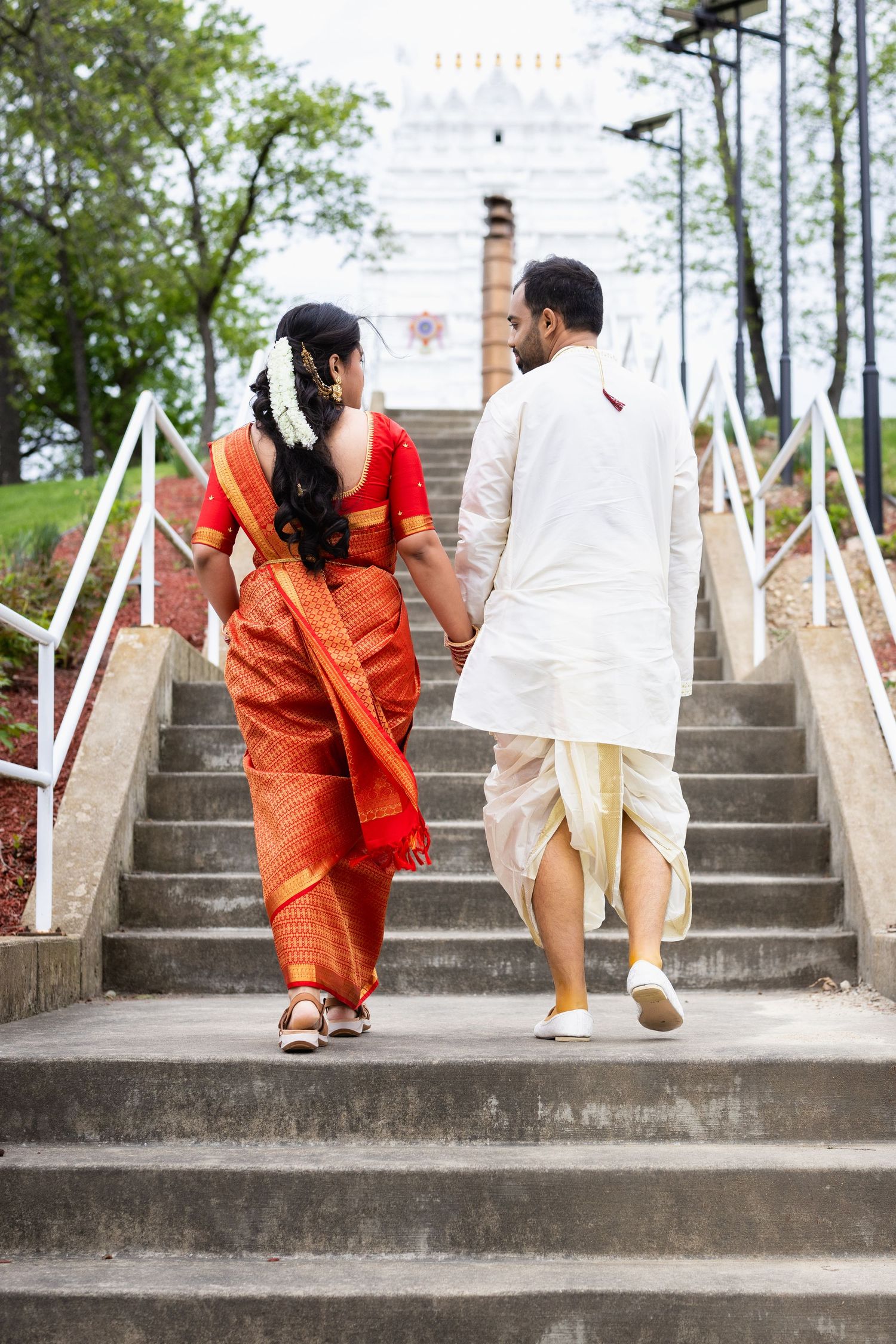 Indian wedding couple climbing stairs hand in hand toward white temple, bride in red saree and groom in white dhoti.