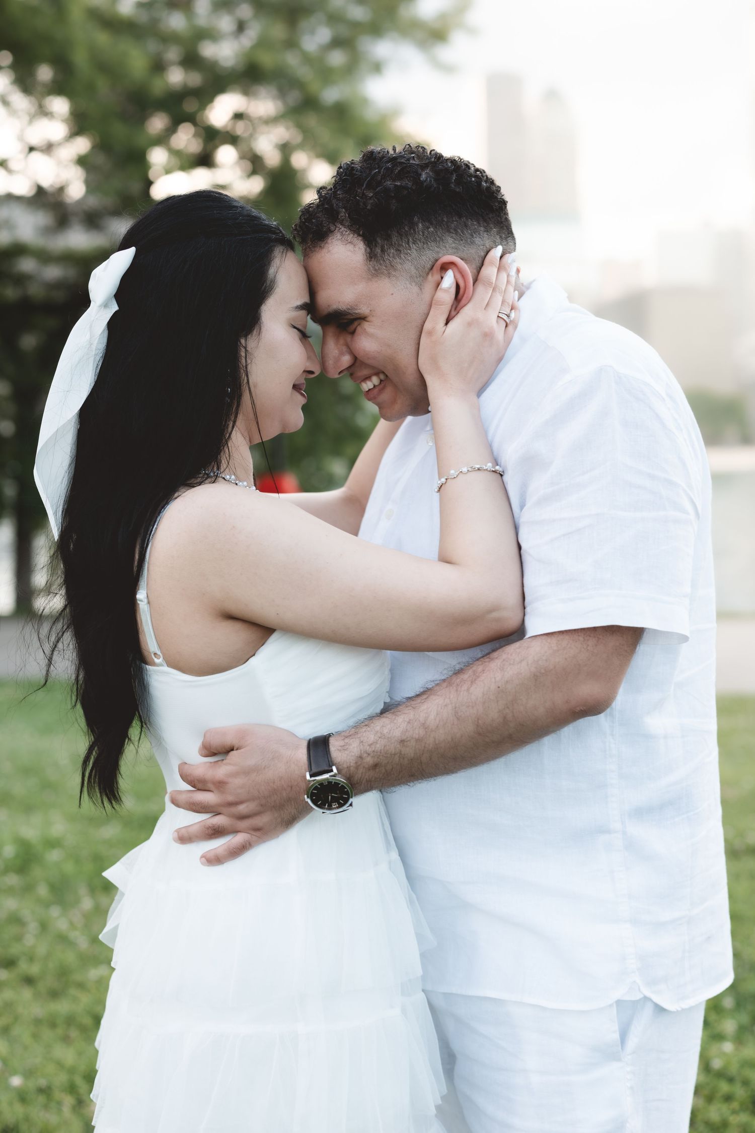 Happy couple in white attire sharing a romantic forehead touch while embracing in a park.