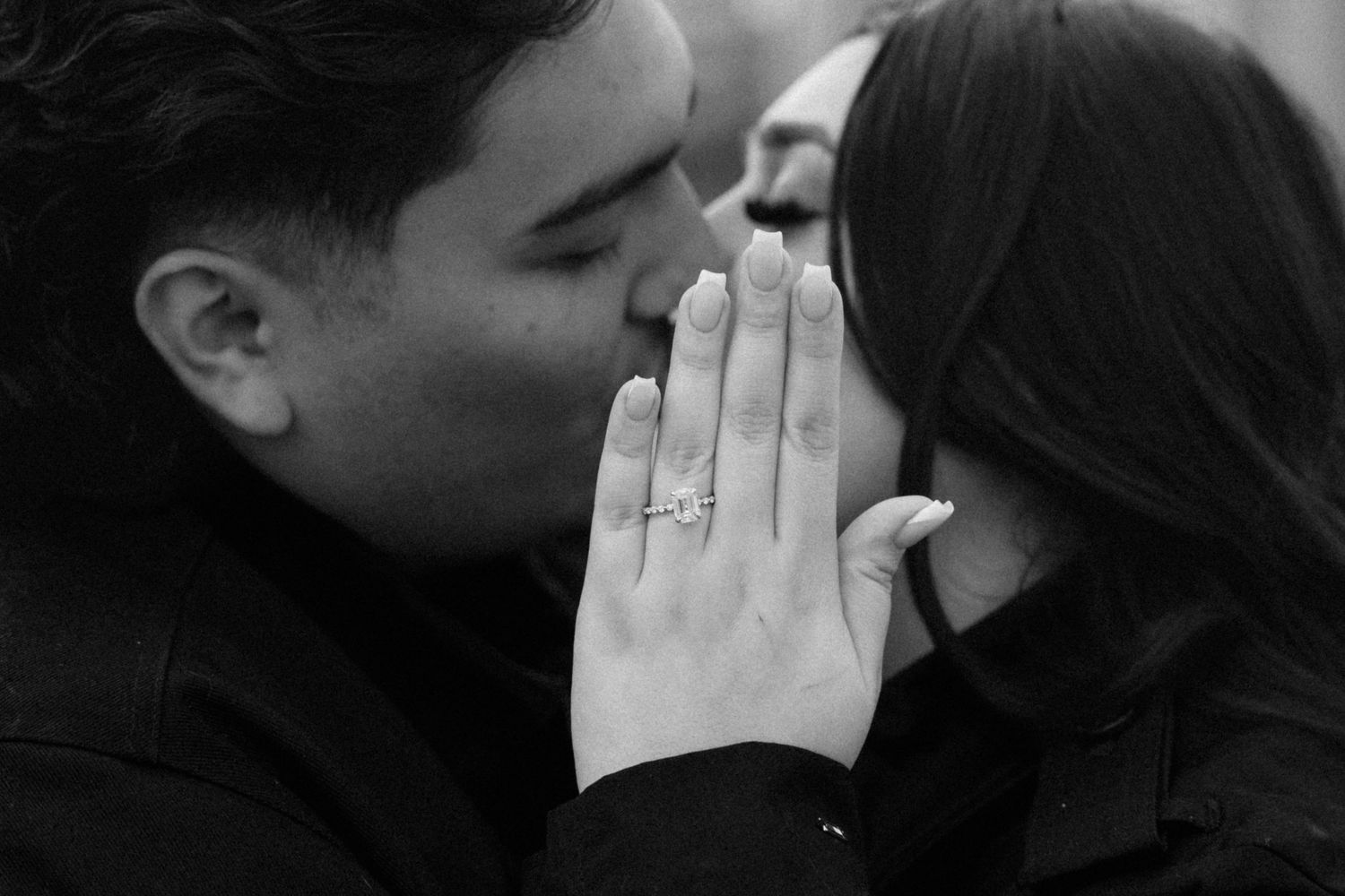 Close-up black and white photo of a couple sharing a kiss with an engagement ring visible on her hand.