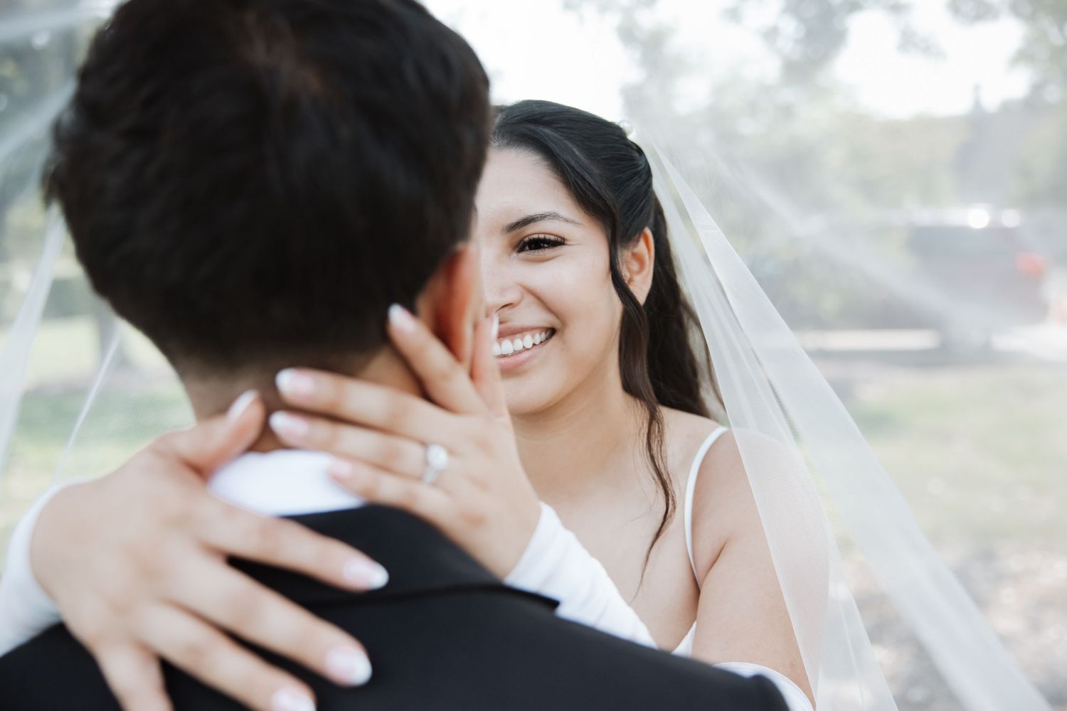 Glowing bride laughs and holds grooms shoulders under a white veil, showcasing her diamond engagement ring.