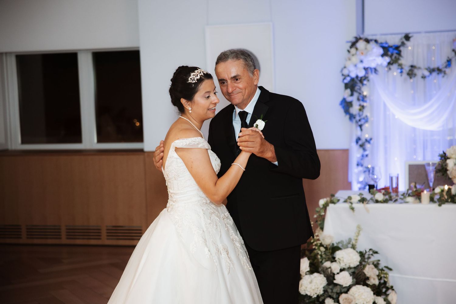 Bride in ball gown and tiara shares father-daughter dance at wedding with floral backdrop.