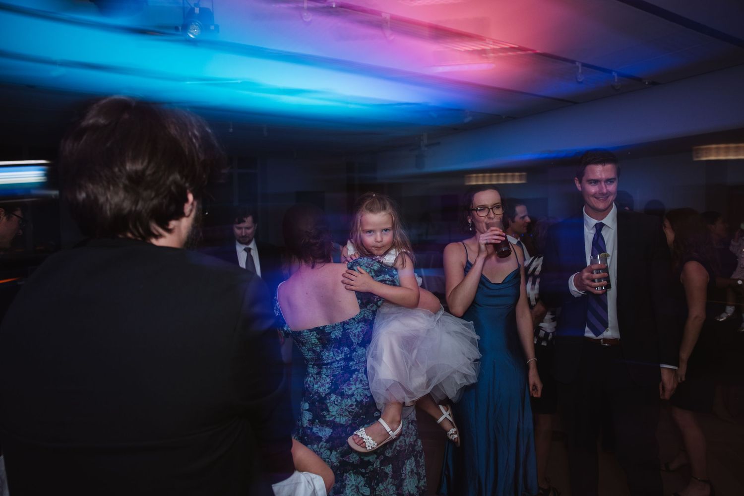 Guests dance and celebrate on a colorfully lit wedding reception dance floor with blue and red lighting.