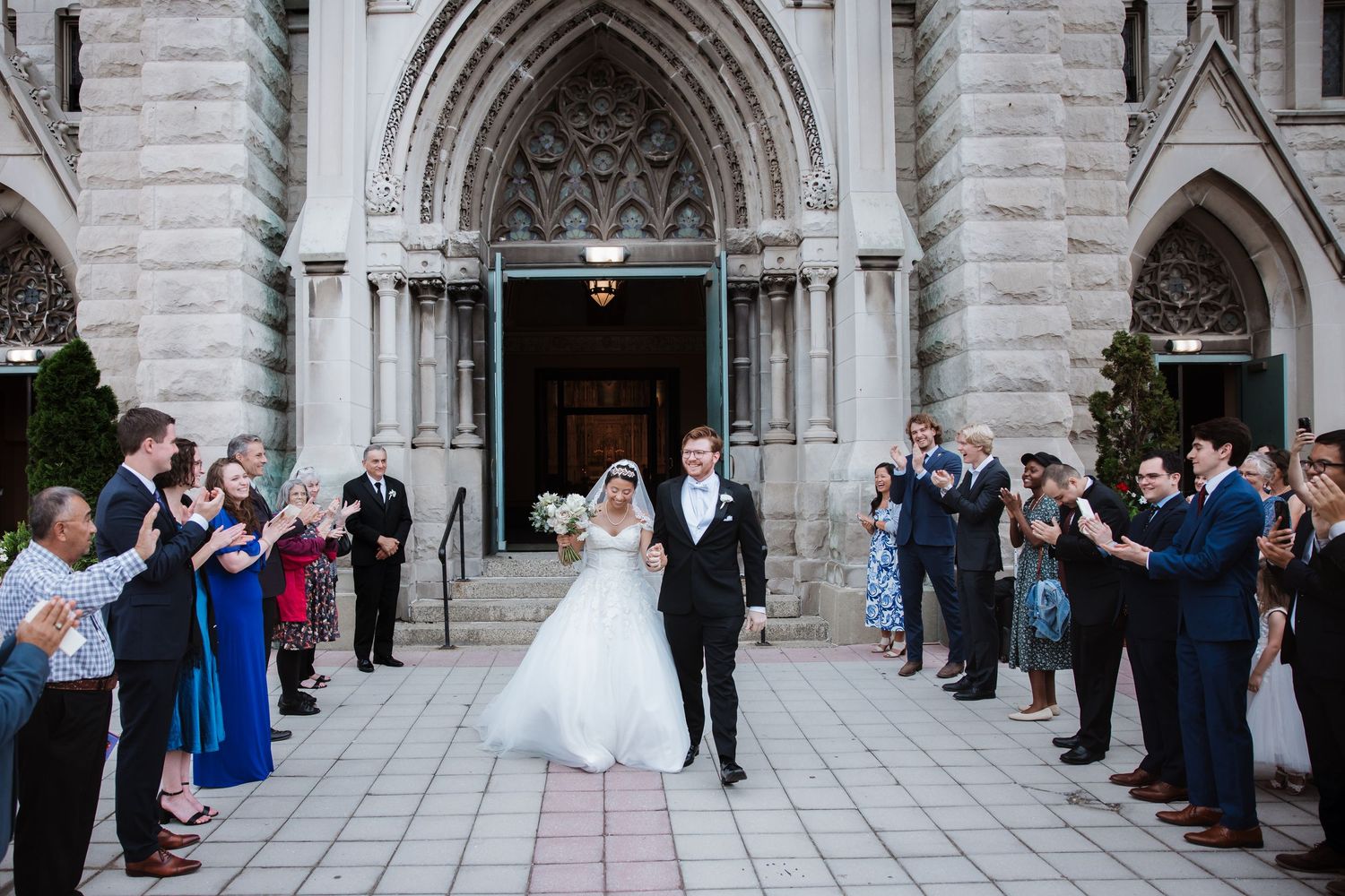 Smiling bride and groom exit grand gothic church entrance surrounded by applauding wedding guests.