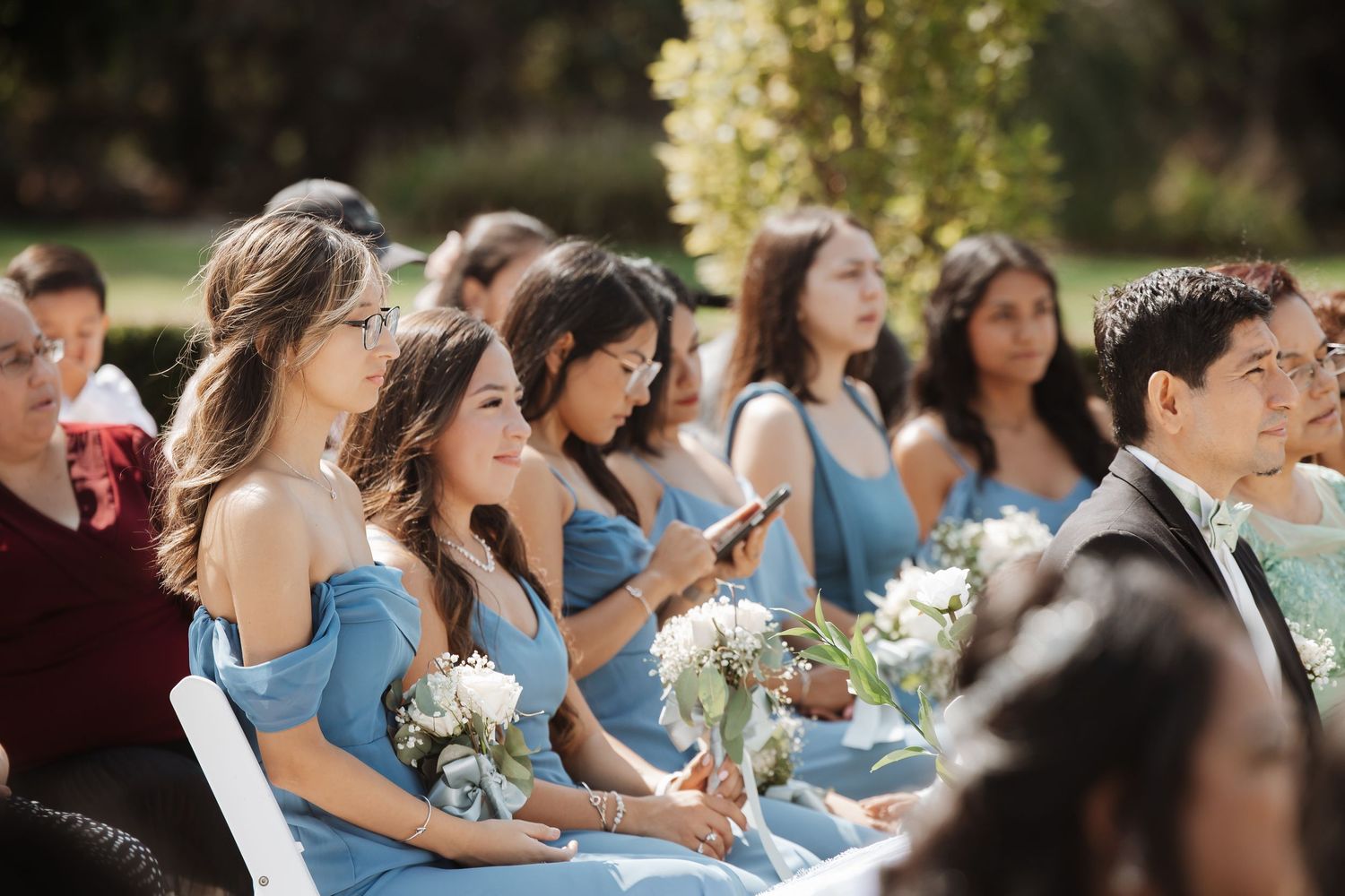 Smiling bridesmaids in off-shoulder blue dresses with white floral bouquets at an outdoor wedding.