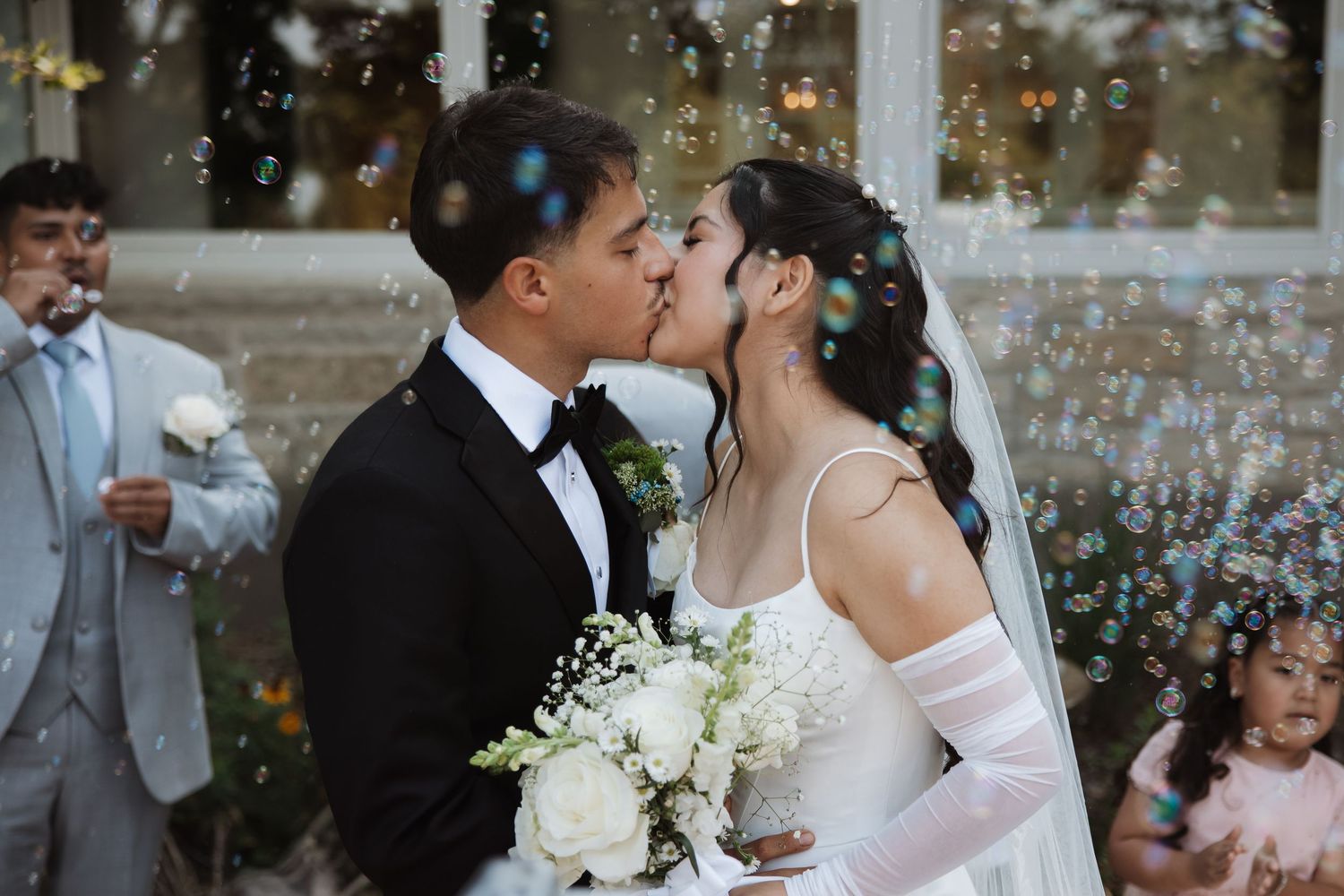 Bride and groom kissing romantically surrounded by floating bubbles with wedding guests in the background.