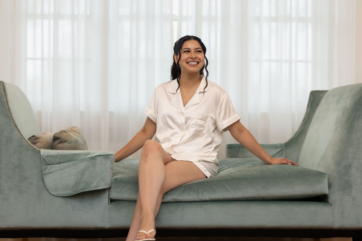 Bride in white satin pajamas relaxing on a sage green velvet sofa before wedding.
