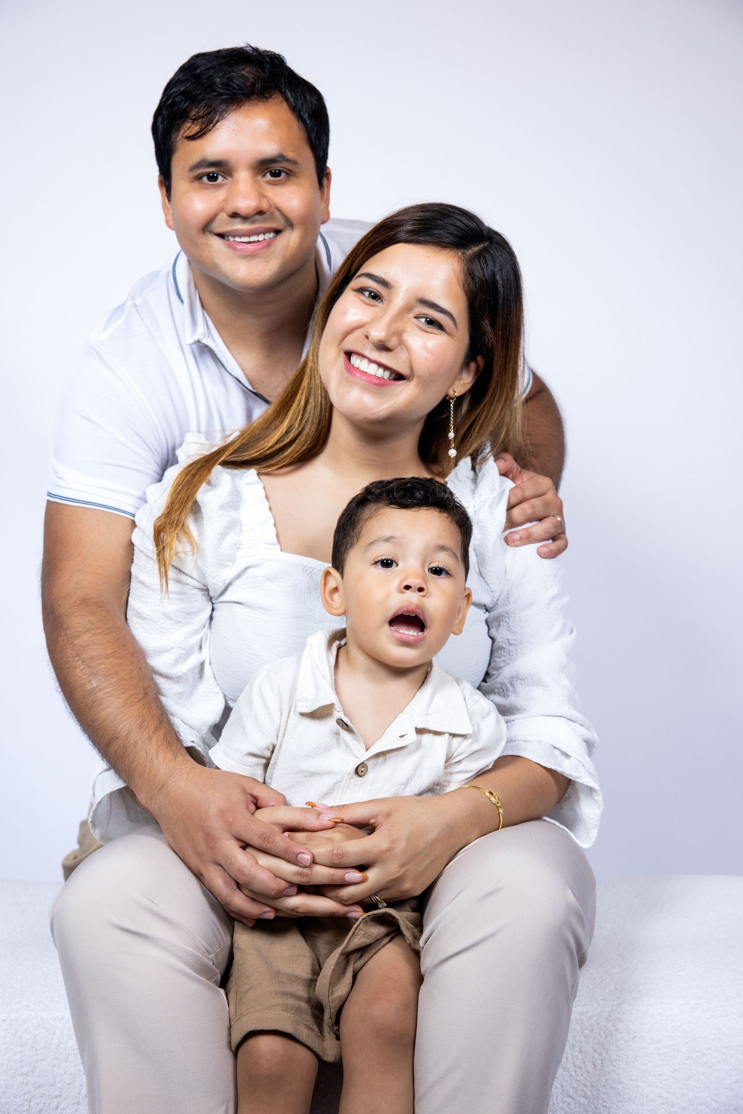 Smiling family portrait with father, mother, and young son in white outfits on white background.