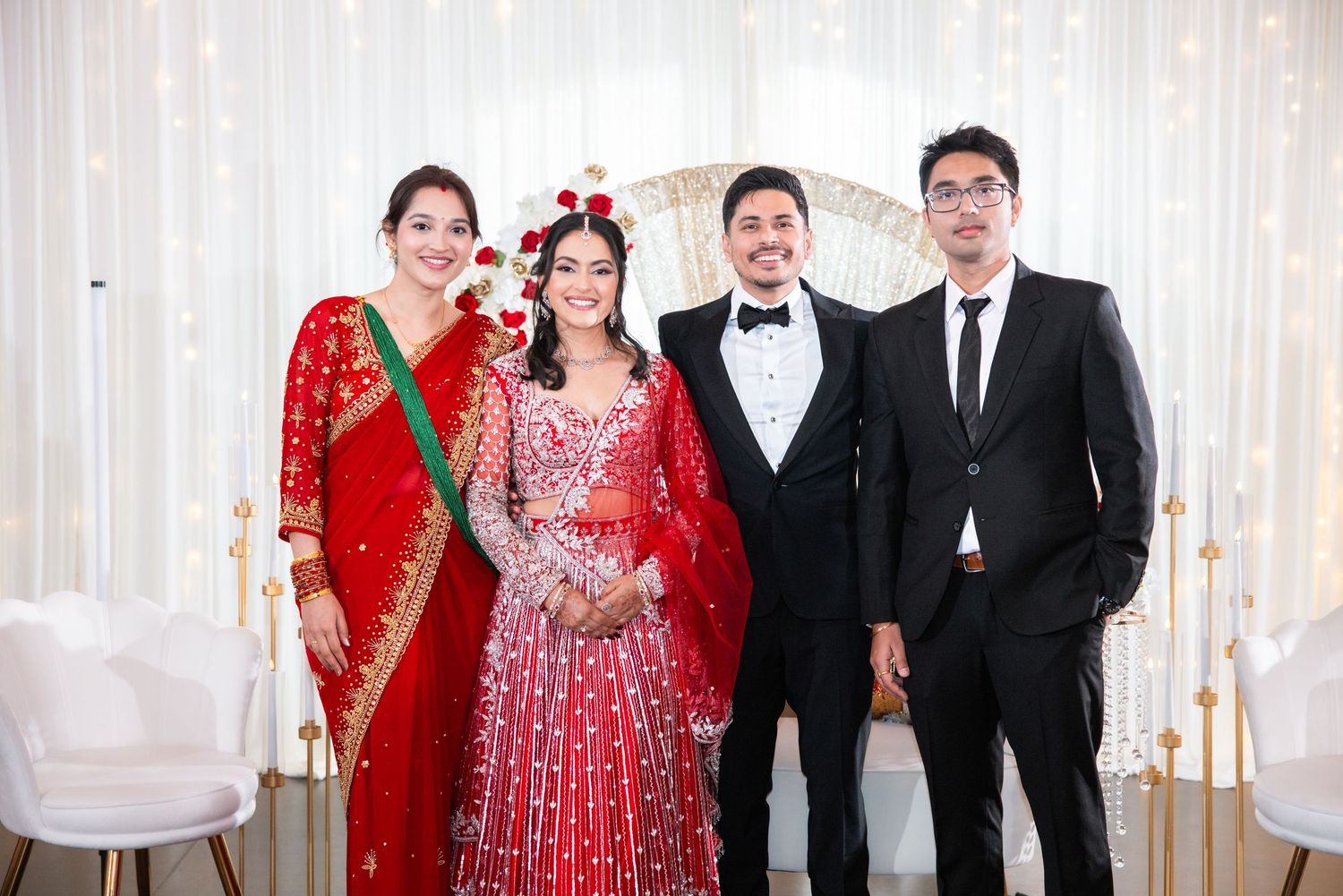 South Asian wedding portrait of bride in red lehenga, groom in tuxedo, posing with woman in red sari and young man.