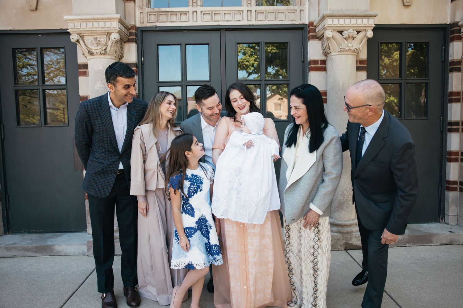 Family group gathered outdoors admiring baby in white christening outfit at baptism celebration.