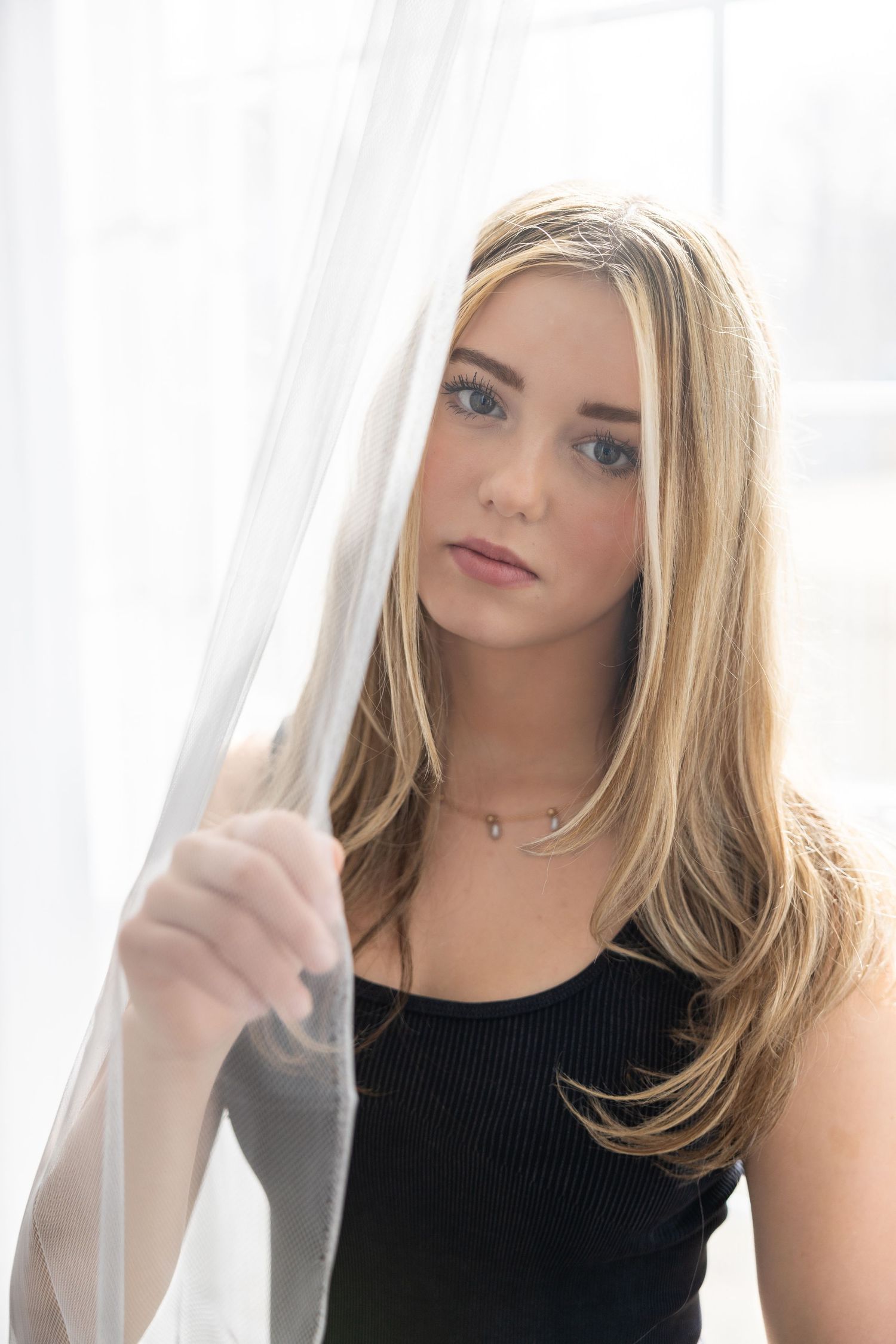 Young blonde woman with highlights peers through sheer white curtain in black tank top indoors.