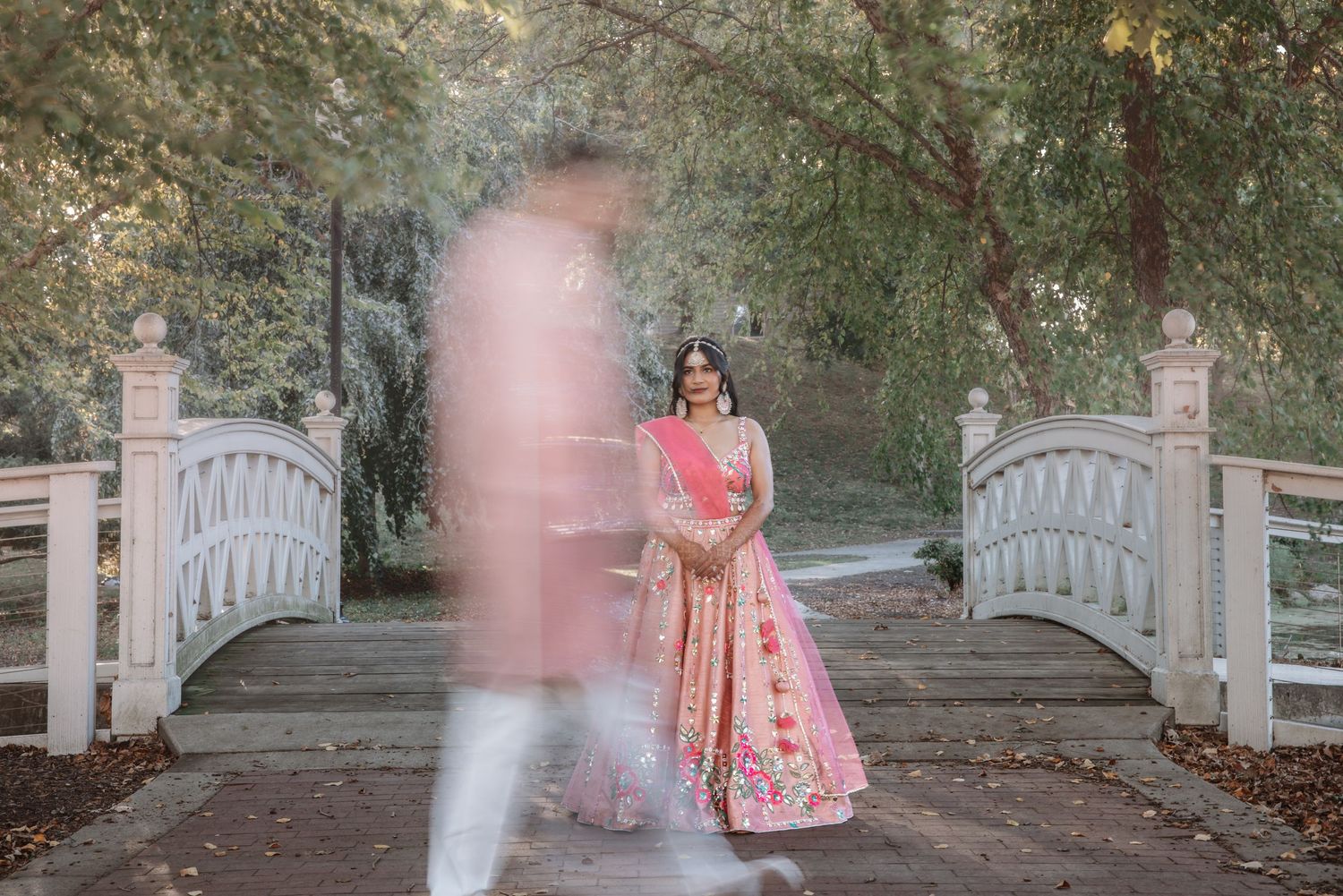 Couple in pink Indian wedding attire on ornate white bridge in lush garden.