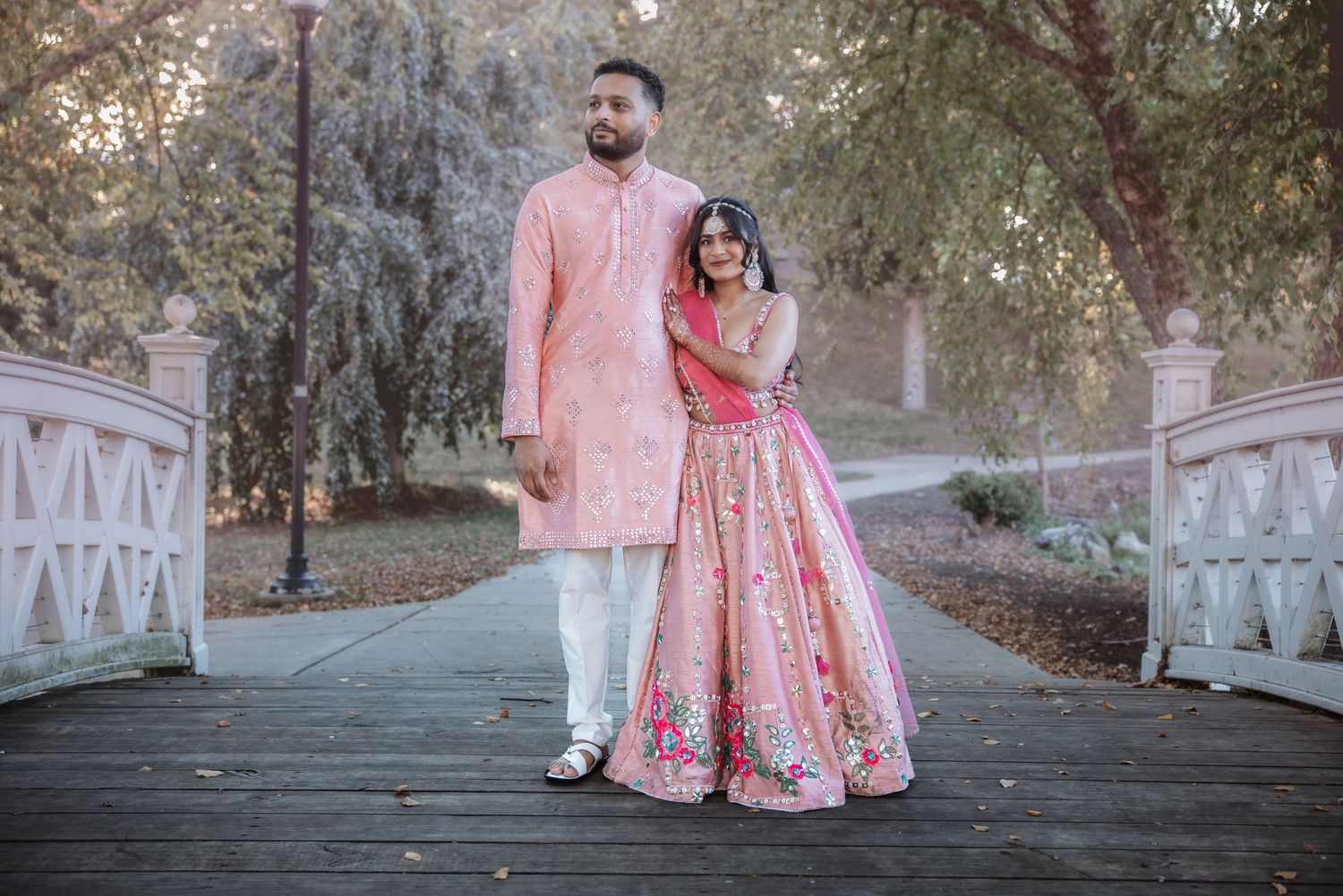 Indian couple in coordinating pink traditional outfits walking on wooden bridge outdoors.